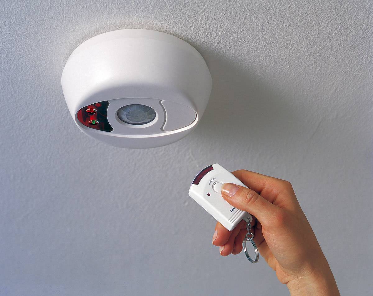 Hand hält Fernbedienung nahe an einer Decken-Sicherheitseinheit. Die Einheit zeigt eine LED-Anzeige und einen Bewegungssensor.