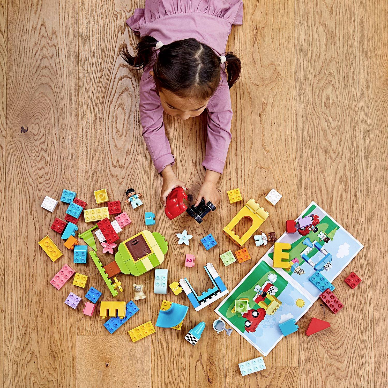Kind spielt mit bunten Bausteinen und Figuren vor einem offenen Bilderbuch auf Holzfußboden, kreativ gestaltete Szene.