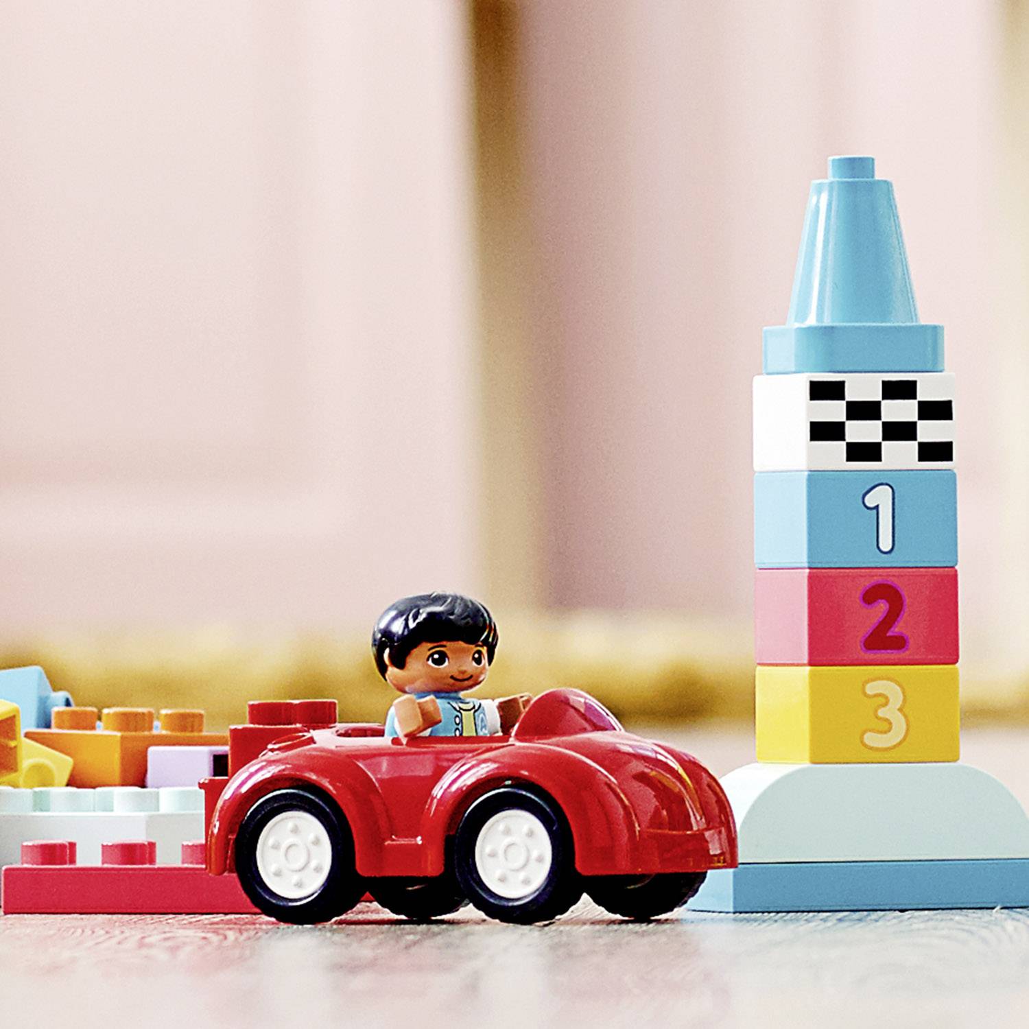 Ein Spielzeugauto mit einer Figur fährt neben einem Zahlen-Turmblock in einem Raum. Der Turm zeigt die Zahlen 1 bis 3.