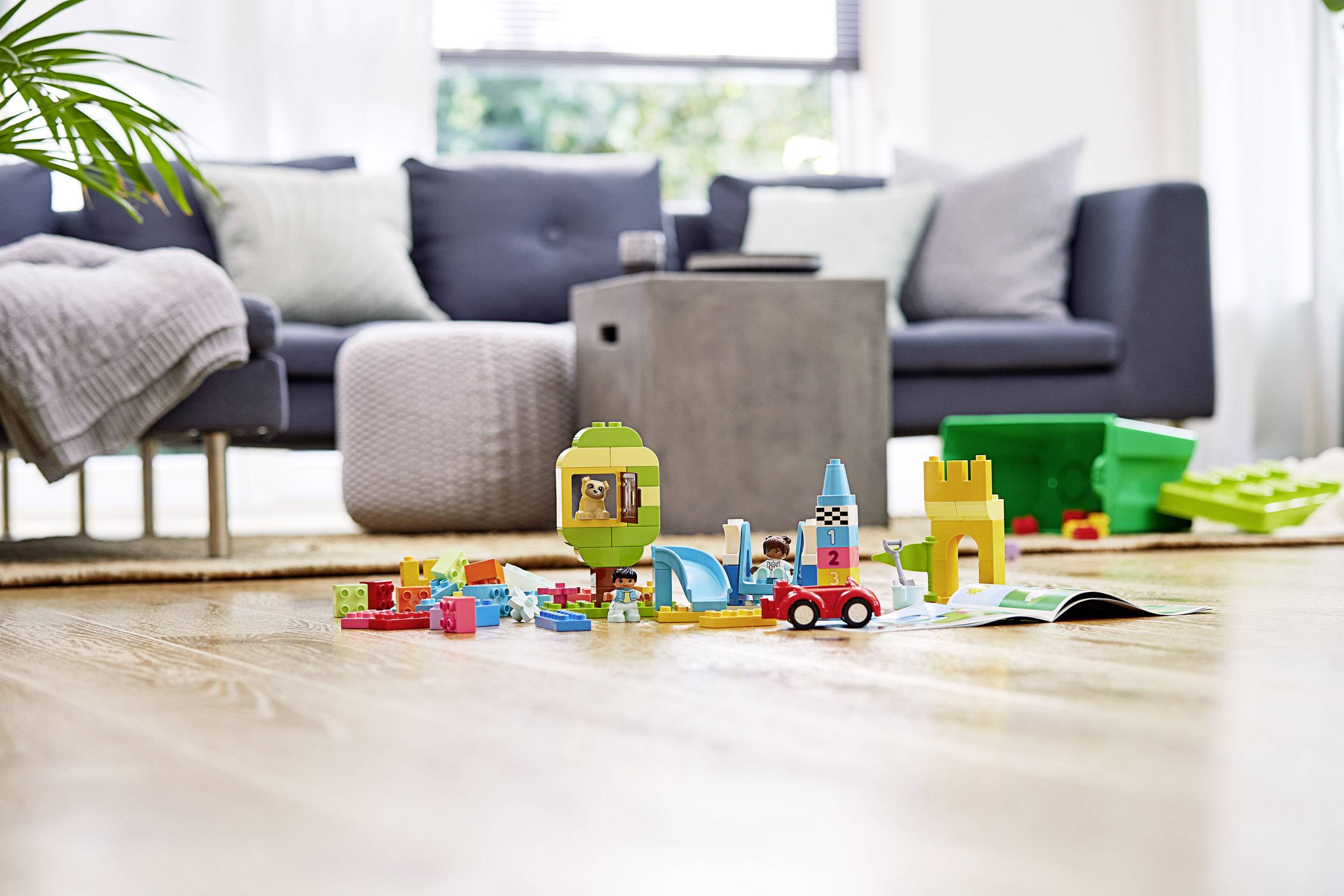 Spielzeugbauklötze und Figuren sind auf dem Boden eines hellen Wohnzimmers verstreut, im Hintergrund ein graues Sofa.