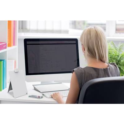 Eine Frau sitzt an einem Schreibtisch und arbeitet an einem Computer. Ein Smartphone liegt daneben. Im Hintergrund sind Pflanzen zu sehen.
