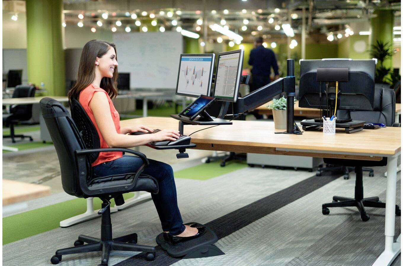 Eine Frau sitzt an einem Schreibtisch in einem Büro und arbeitet mit zwei Computermonitoren. Die Umgebung ist modern und im Open-Space-Design gehalten, mit hellem Licht.