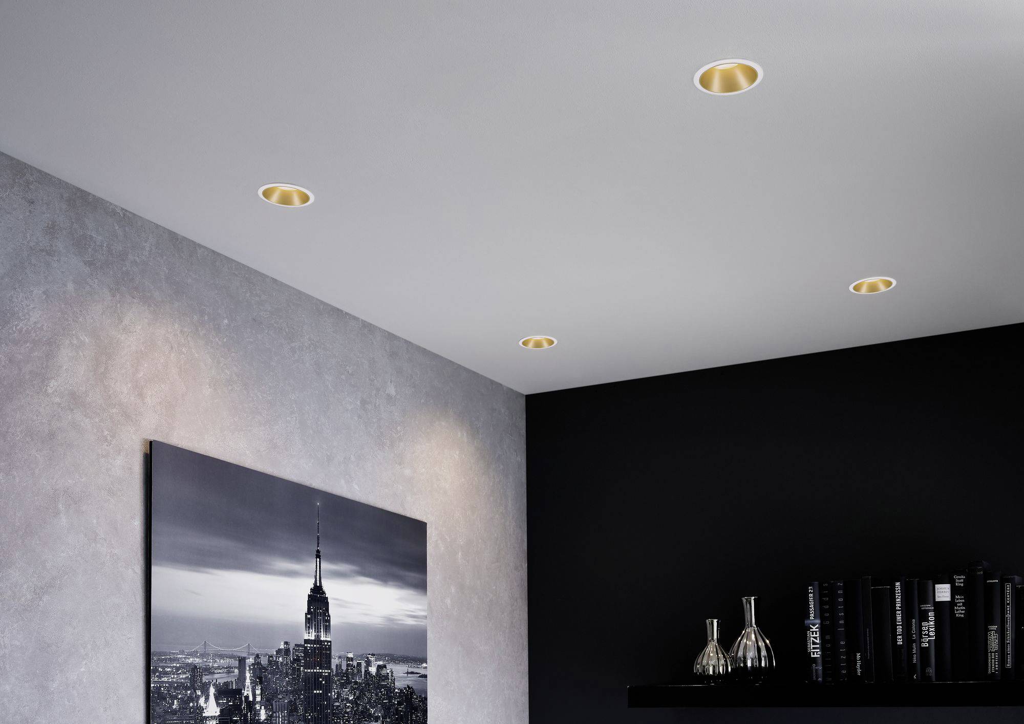 Vier eingelassene Deckenstrahler beleuchten ein Schlafzimmer. An der Wand hängt ein Schwarz-Weiß-Foto einer Stadt. Ein Regal mit Dekorobjekten ist sichtbar.