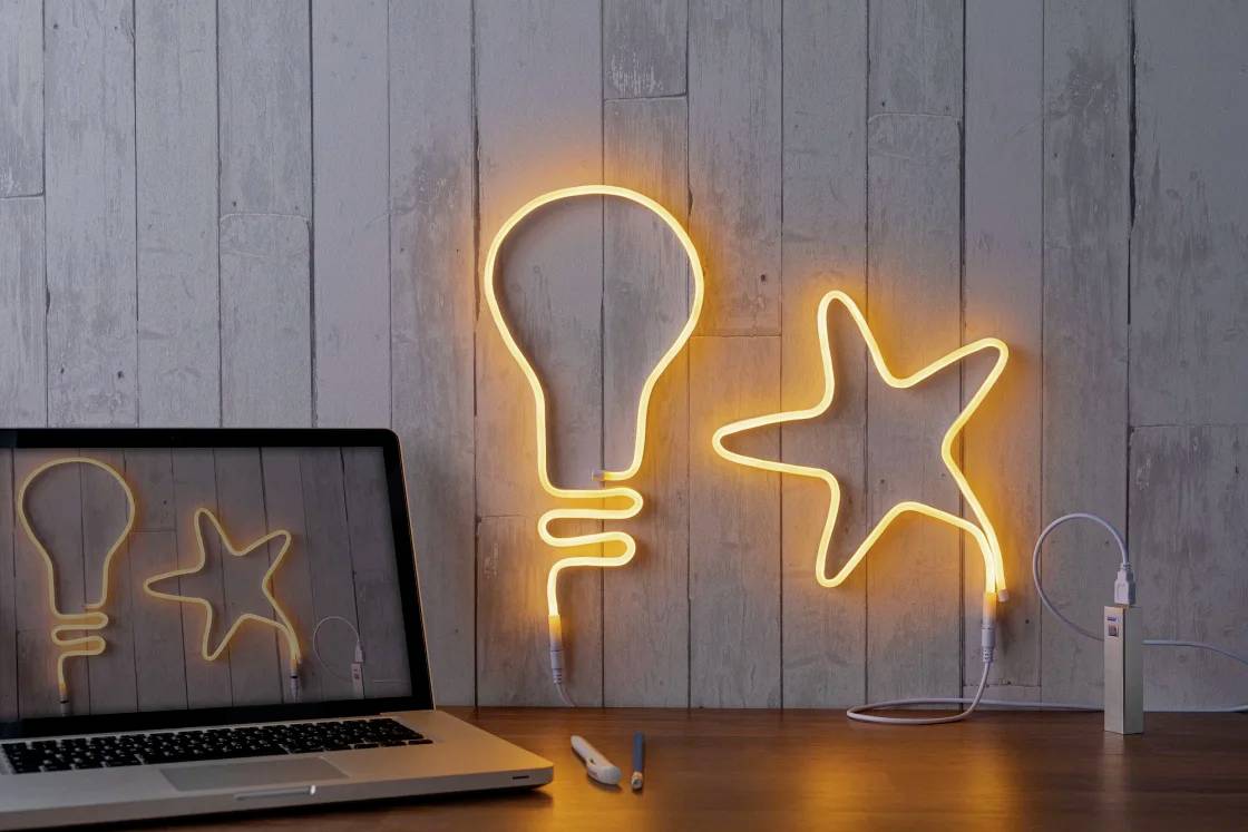 Ein Laptop spiegelt ein orangefarbenes Neonlicht in Form einer Glühbirne und eines Sterns wider, die an einer Holzwand leuchten.