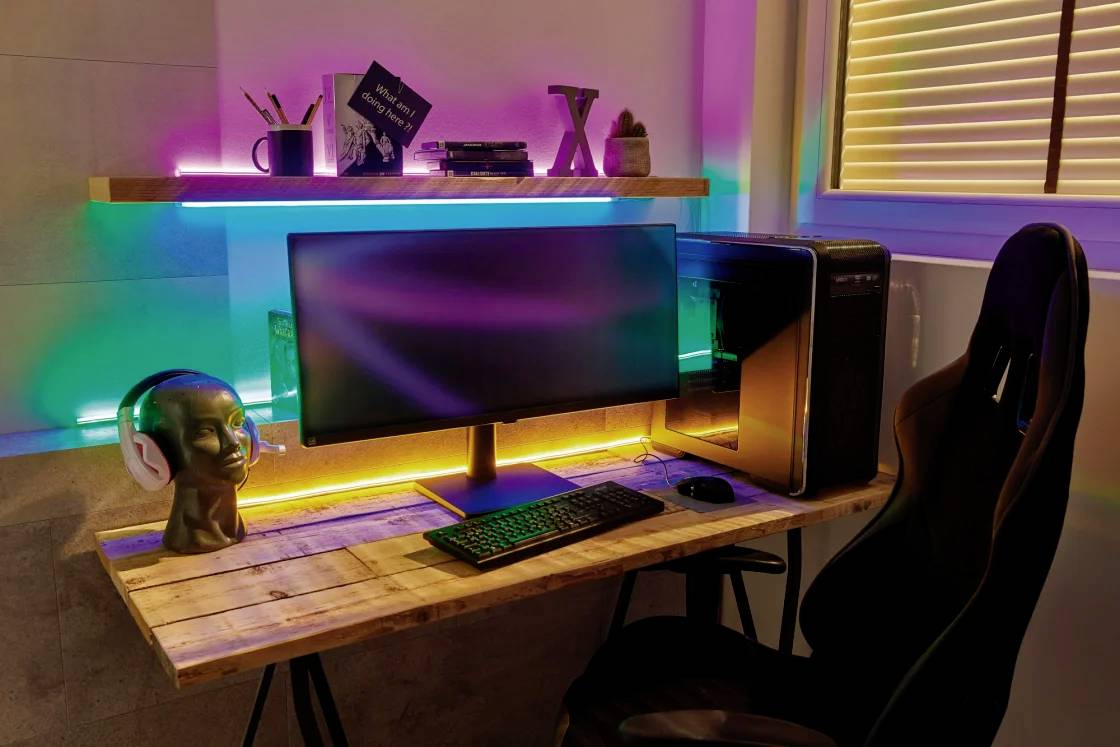 Ein moderner Schreibtisch mit Computer, Monitor, Tastatur und Maus. Bunte LED-Leuchten beleuchten den Raum. Ein Kopfhörerständer steht links.