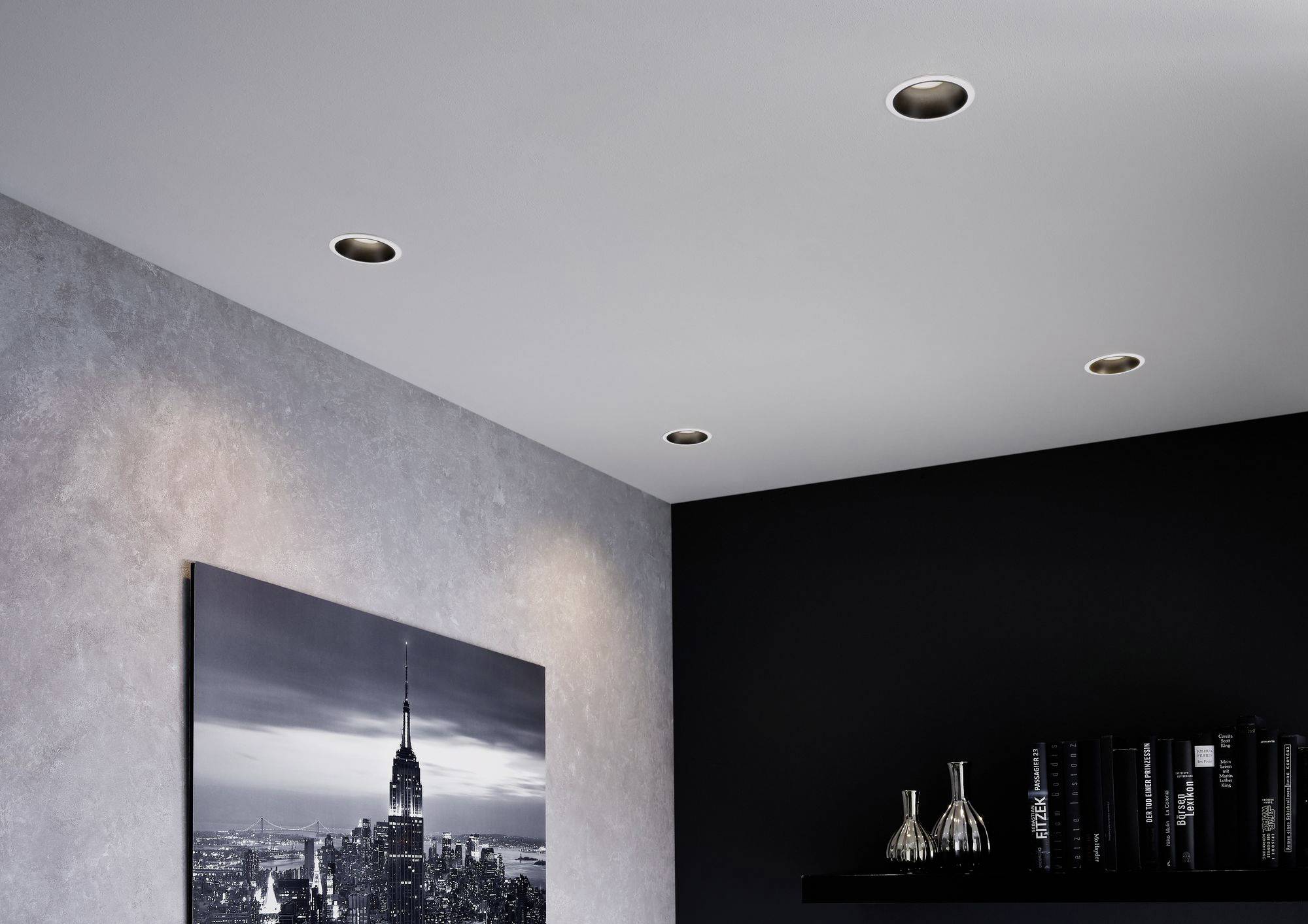 Zimmerdecke mit vier eingebauten Deckenleuchten. An der Wand hängt ein Schwarz-Weiß-Bild einer Stadtlandschaft. Ein Regal mit Vasen und Büchern steht darunter.