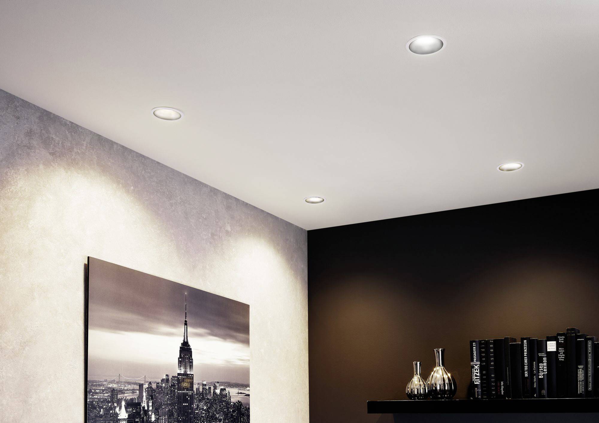 Deckenbeleuchtung mit vier eingebauten Spots beleuchtet ein Wohnzimmer mit einem Schwarz-Weiß-Bild und schwarzen Büchern auf einem Regal.