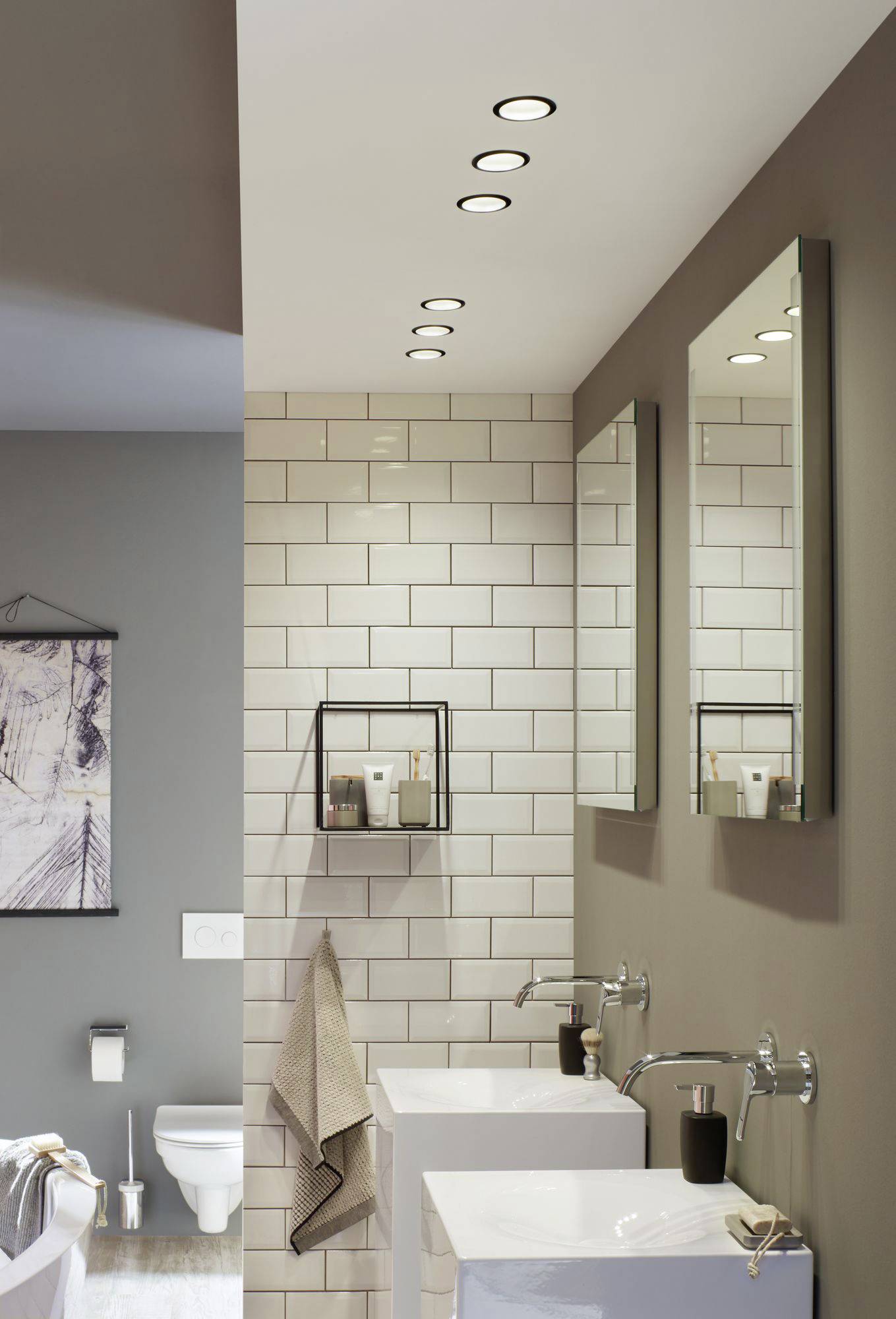 Modernes Badezimmer mit weißen Fliesen, zwei Waschbecken mit Spiegeln, Wandregal mit Pflegeprodukten und hinterer Blick auf die Toilette.