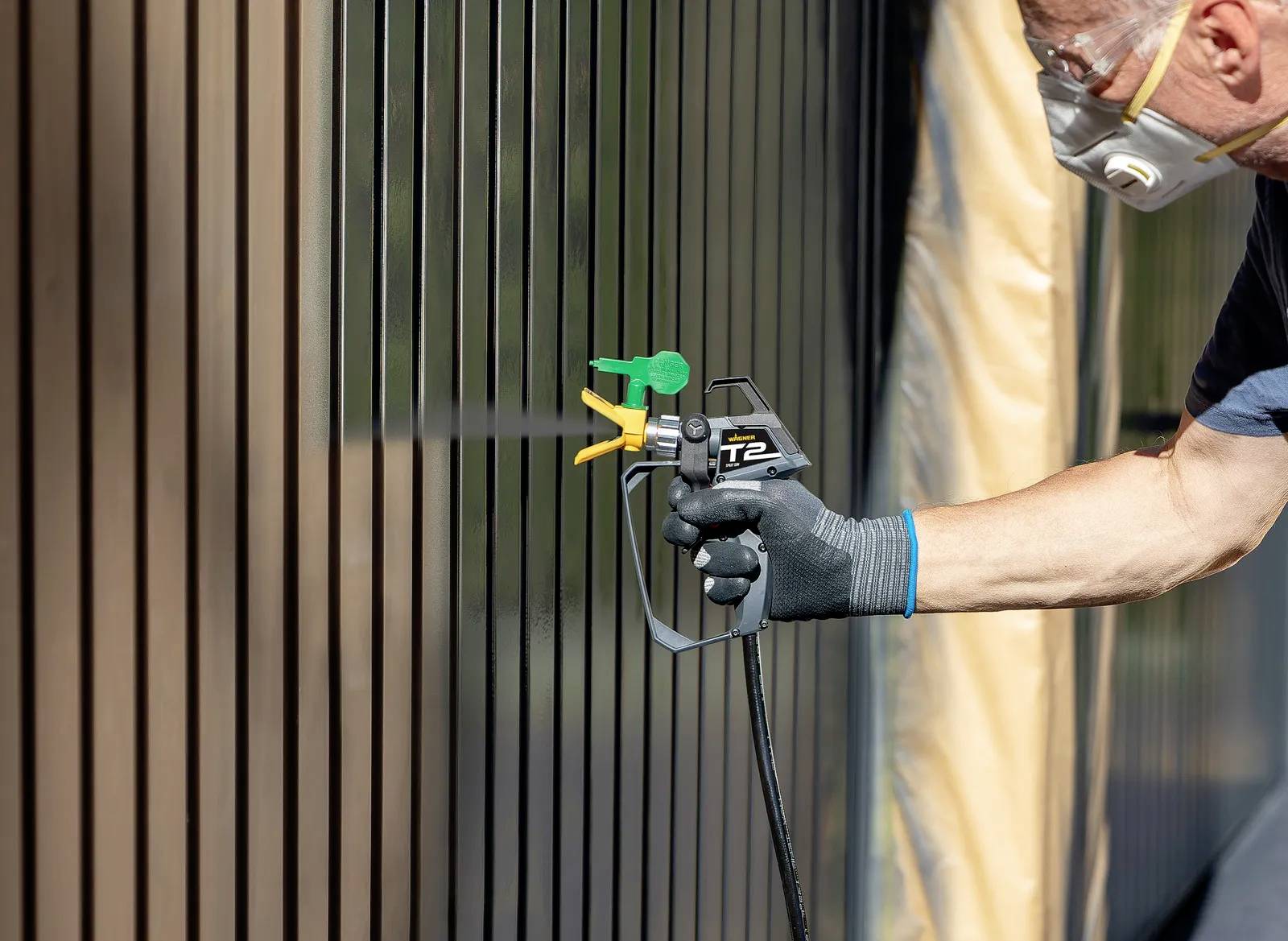 Eine Person sprüht mit einer Lackierpistole Farbe auf eine Metallwand. Sie trägt eine Schutzmaske und Handschuhe.