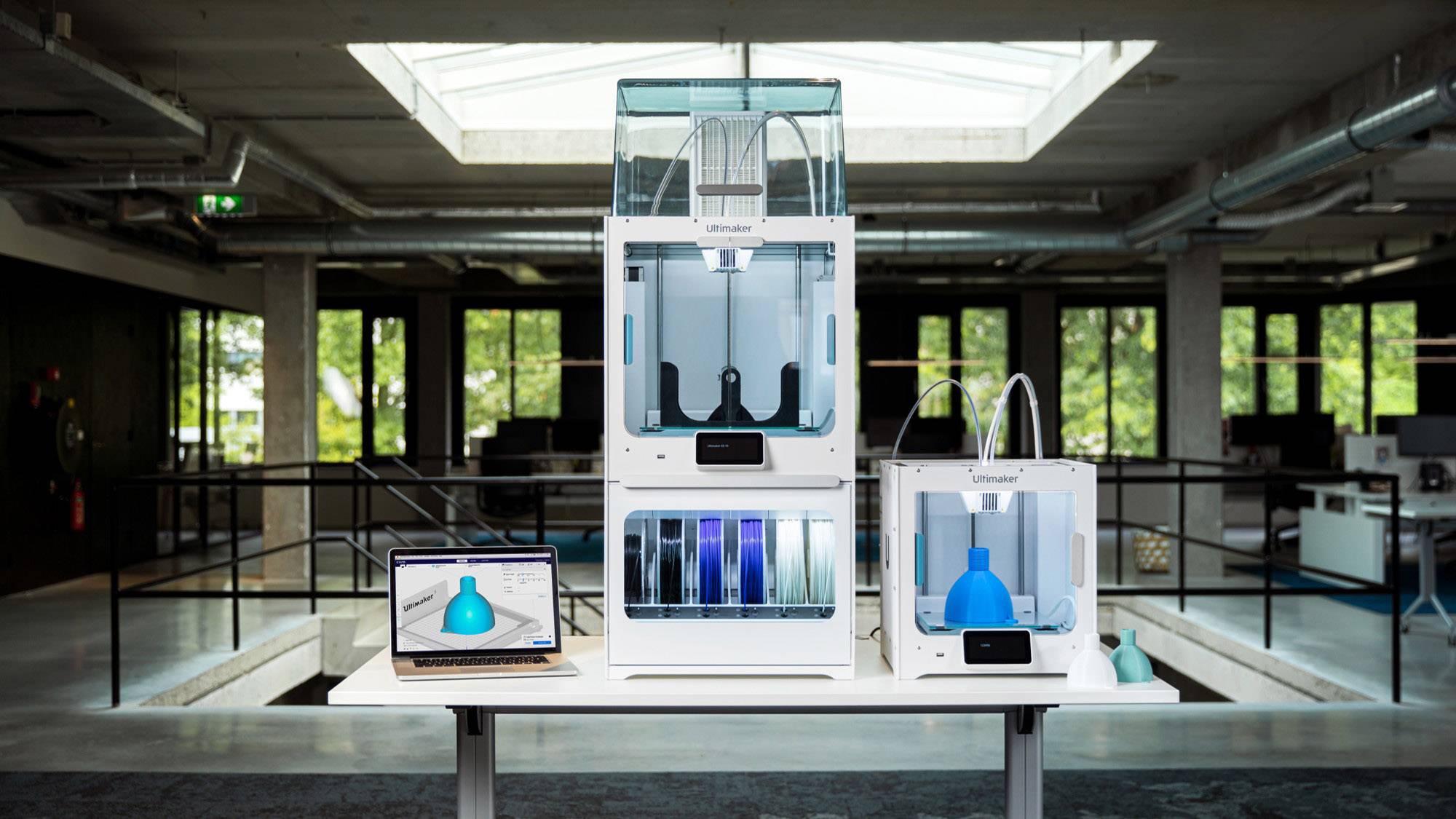 Ein moderner Büroraum mit einem Tisch, auf dem ein Laptop und mehrere 3D-Drucker stehen. Der Laptop zeigt ein blaues 3D-Modell.