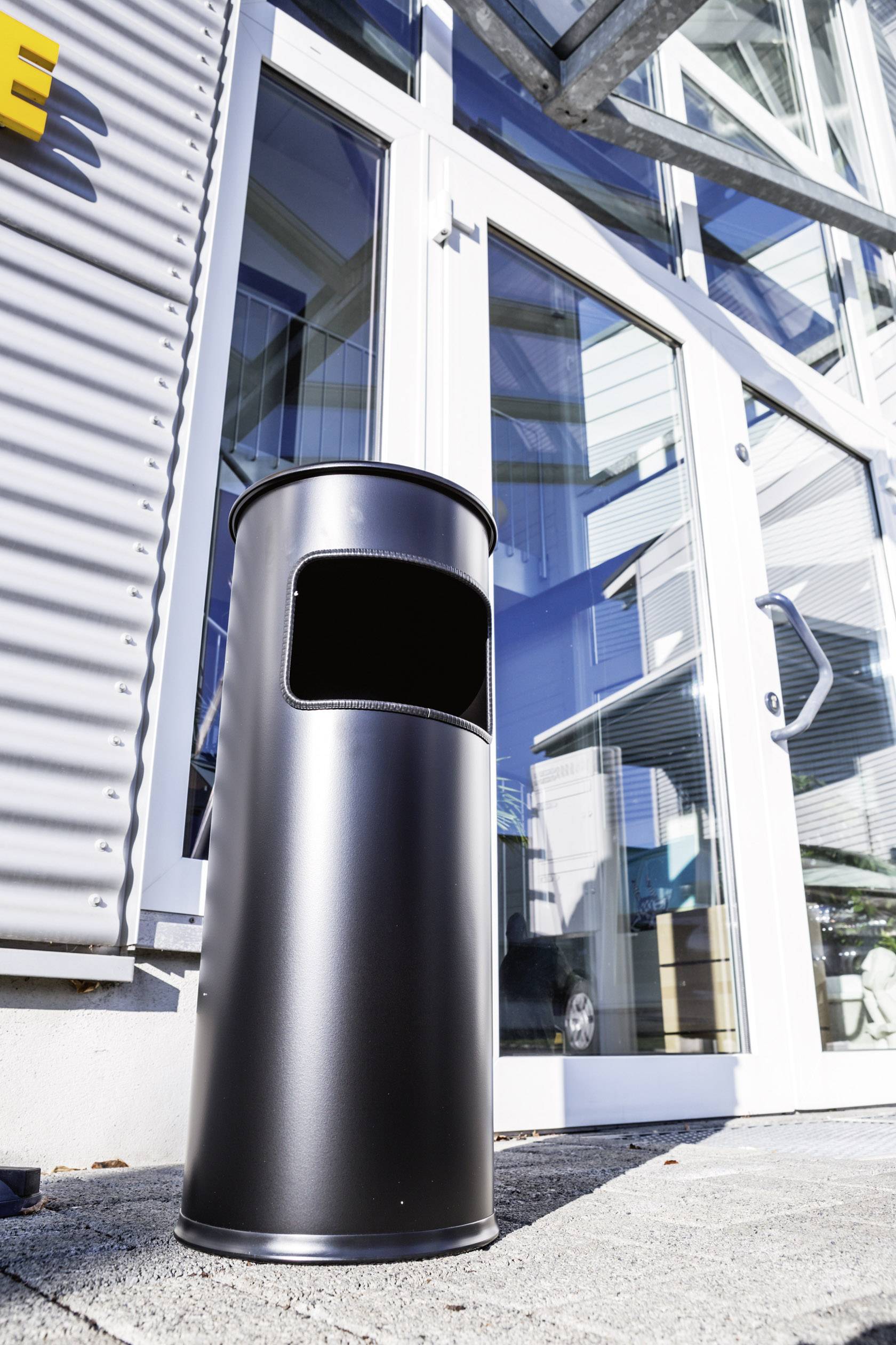 Ein runder, schwarzer Abfalleimer steht vor einem gläsernen Eingang, der teilweise ein Auto im Inneren eines Gebäudes zeigt.