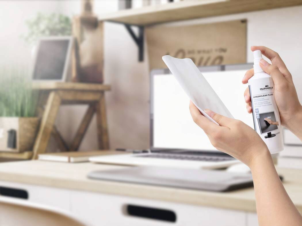 Eine Person, die einen Tablet-Bildschirm mit einer Sprühflasche in einem Home-Office-Umfeld reinigt, mit einem Laptop, einer Pflanze und Regalen im Hintergrund.