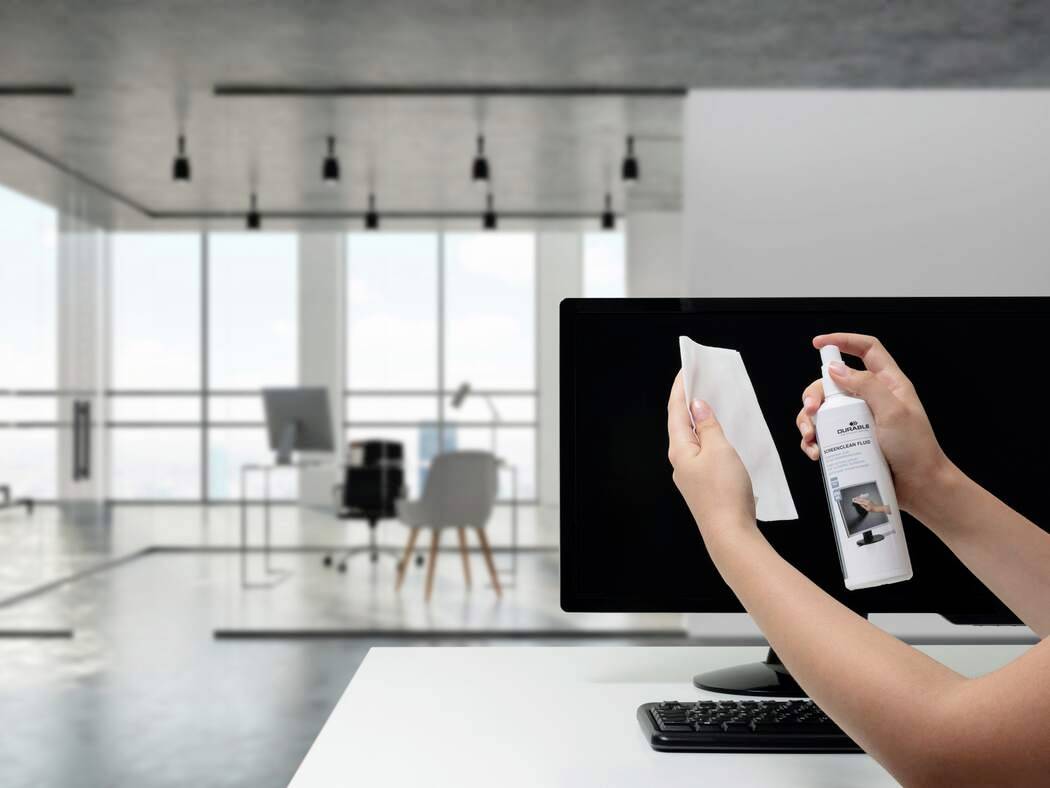 Eine Hand hält eine Sprühflasche mit der Aufschrift „Bildschirmreiniger" und ein Tuch reinigt einen Computerbildschirm. Ein moderner Büroraum befindet sich im Hintergrund.