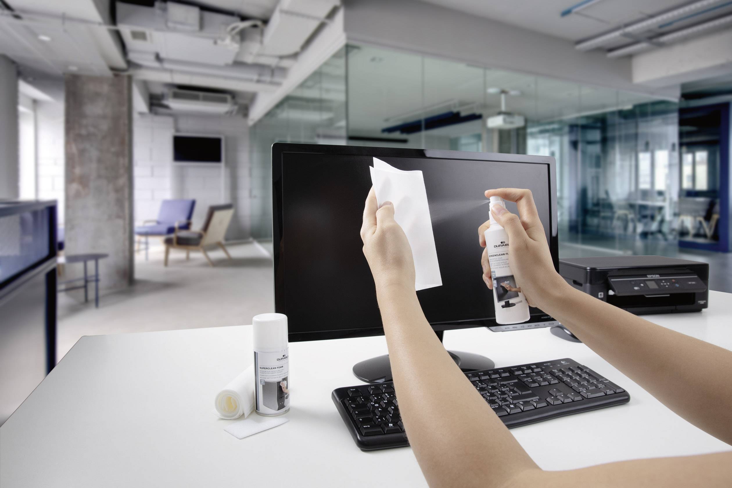 Eine Person reinigt einen Computermonitor mit einem Tuch und Sprühreiniger in einem modernen Büro mit mehreren Arbeitsplätzen.