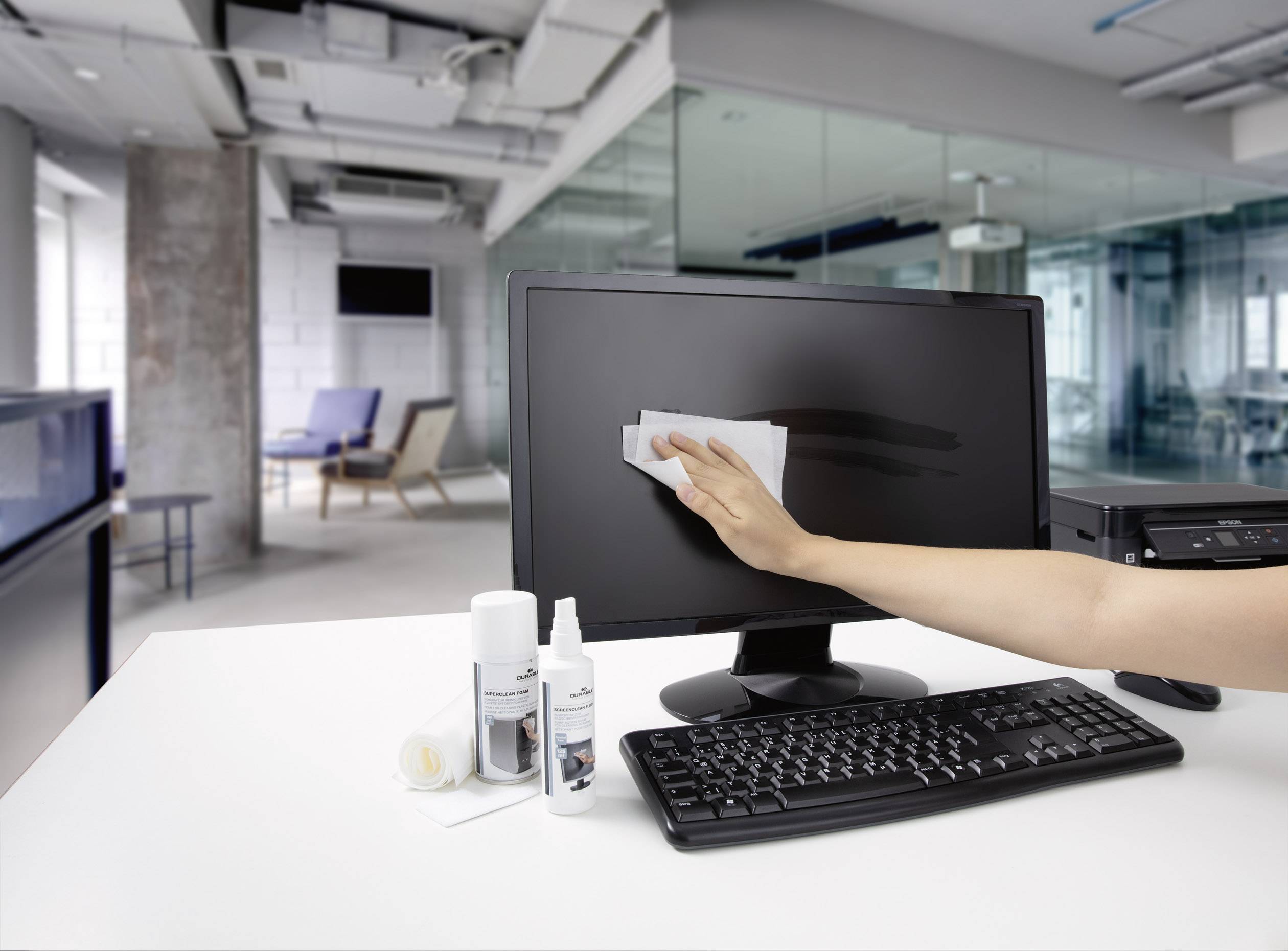 Eine Hand reinigt mit einem Tuch den Bildschirm eines Computers in einem modernen Büro. Tisch mit Tastatur, Monitor, Reinigungsmitteln.