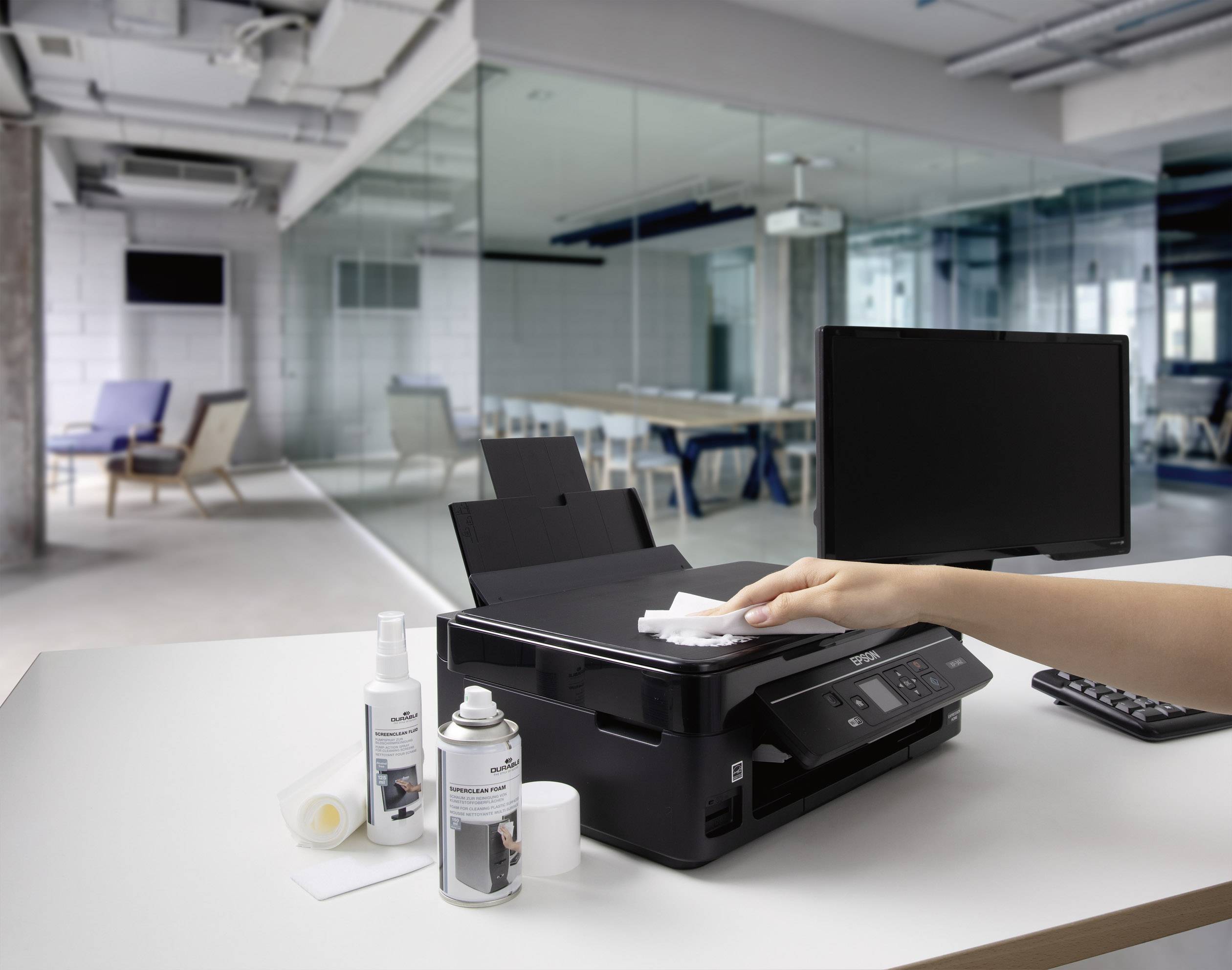 'Eine Person reinigt einen Drucker in einem modernen Büro. Neben dem Drucker stehen Reinigungsprodukte, ein Computerbildschirm und eine Tastatur.'