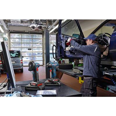 Ein Mechaniker in einer Werkstatt repariert das Armaturenbrett eines Autos. Zwei Akkuschrauber stehen im Vordergrund auf einem Tisch.