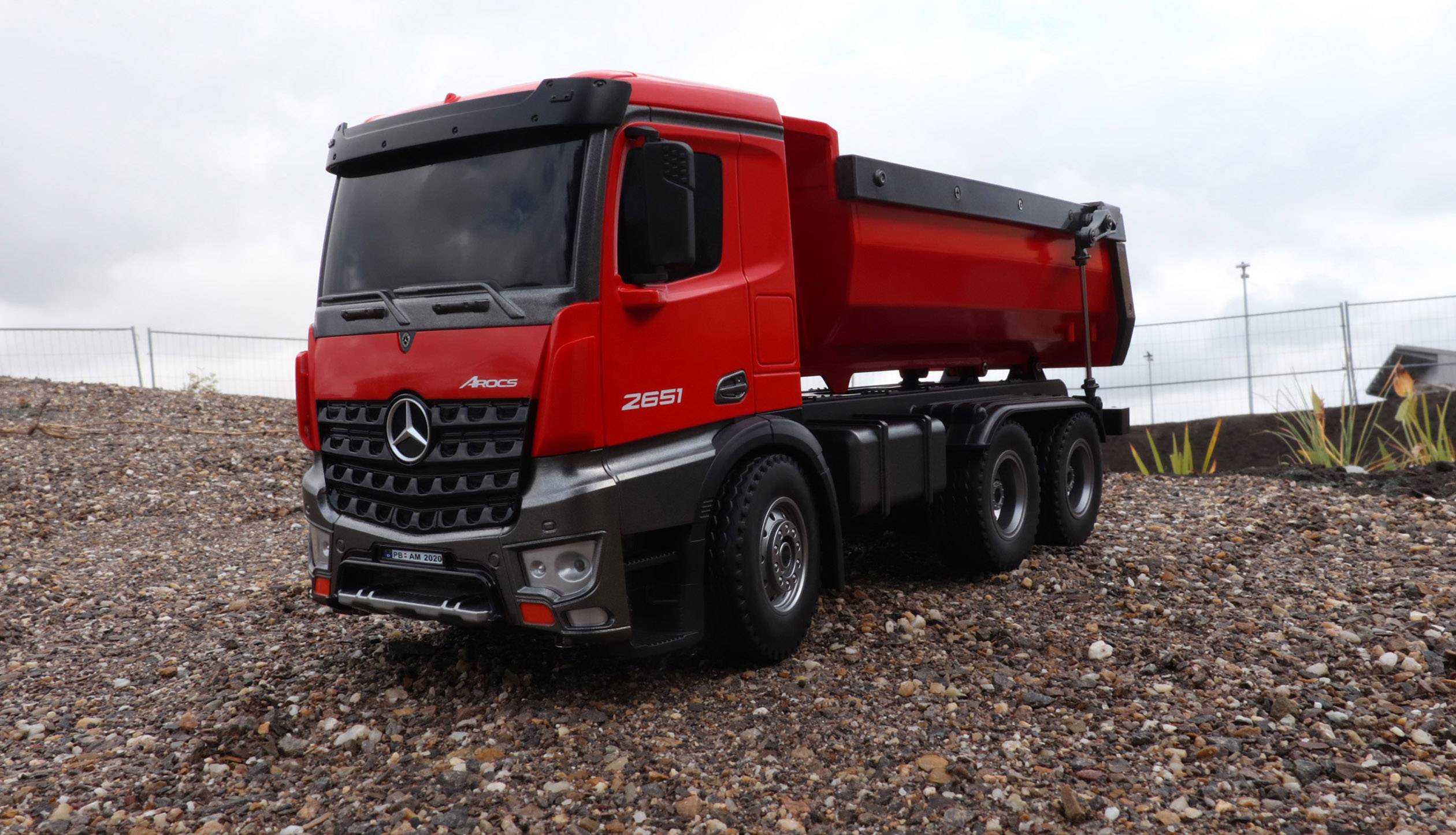 Ein roter, detaillierter Modell-Lkw steht auf einer Kiesfläche. Der Himmel ist bewölkt, und im Hintergrund sind Zäune sichtbar.