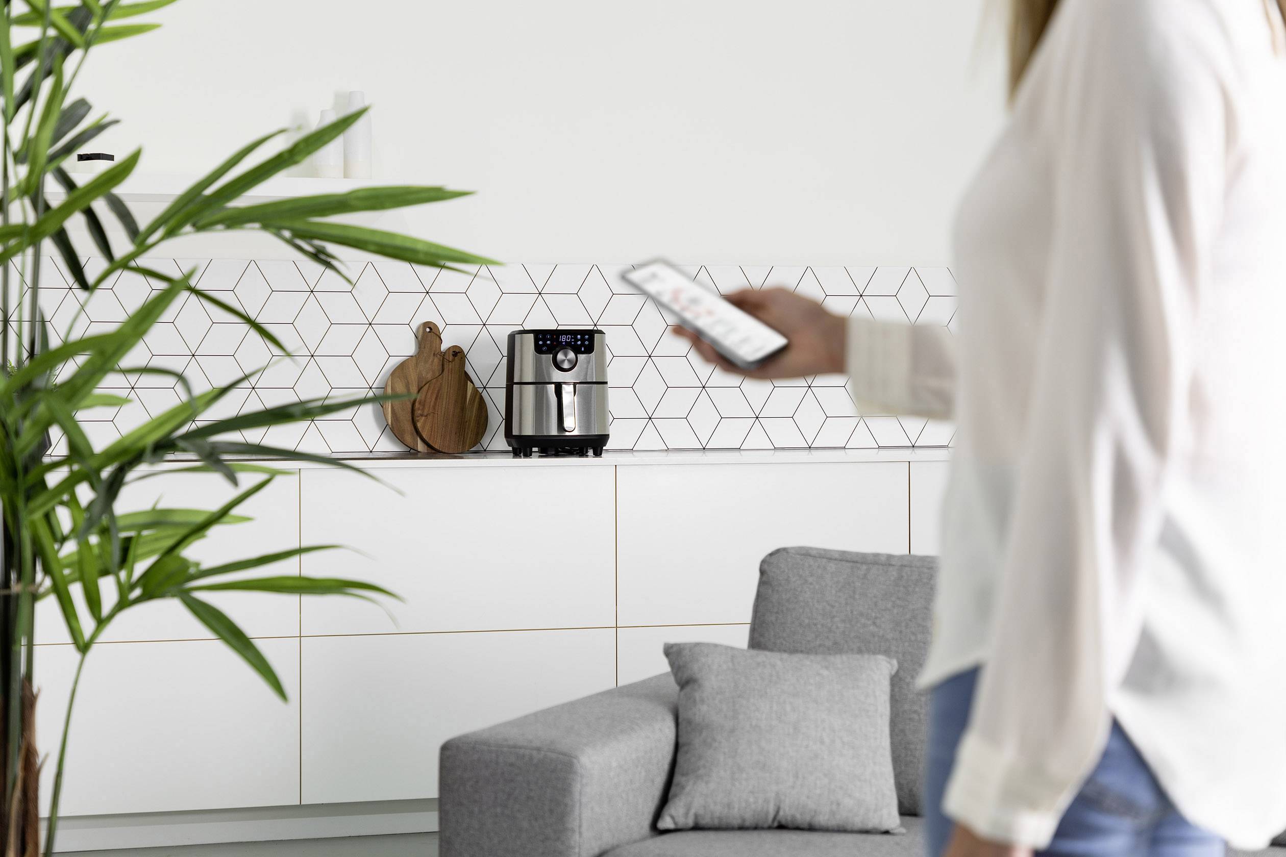 Eine Person hält ein Smartphone und steht vor einer modernen Küche mit weißen Schränken und geometrischen Fliesen. Im Hintergrund ist eine Heißluftfritteuse auf der Arbeitsfläche zu sehen.