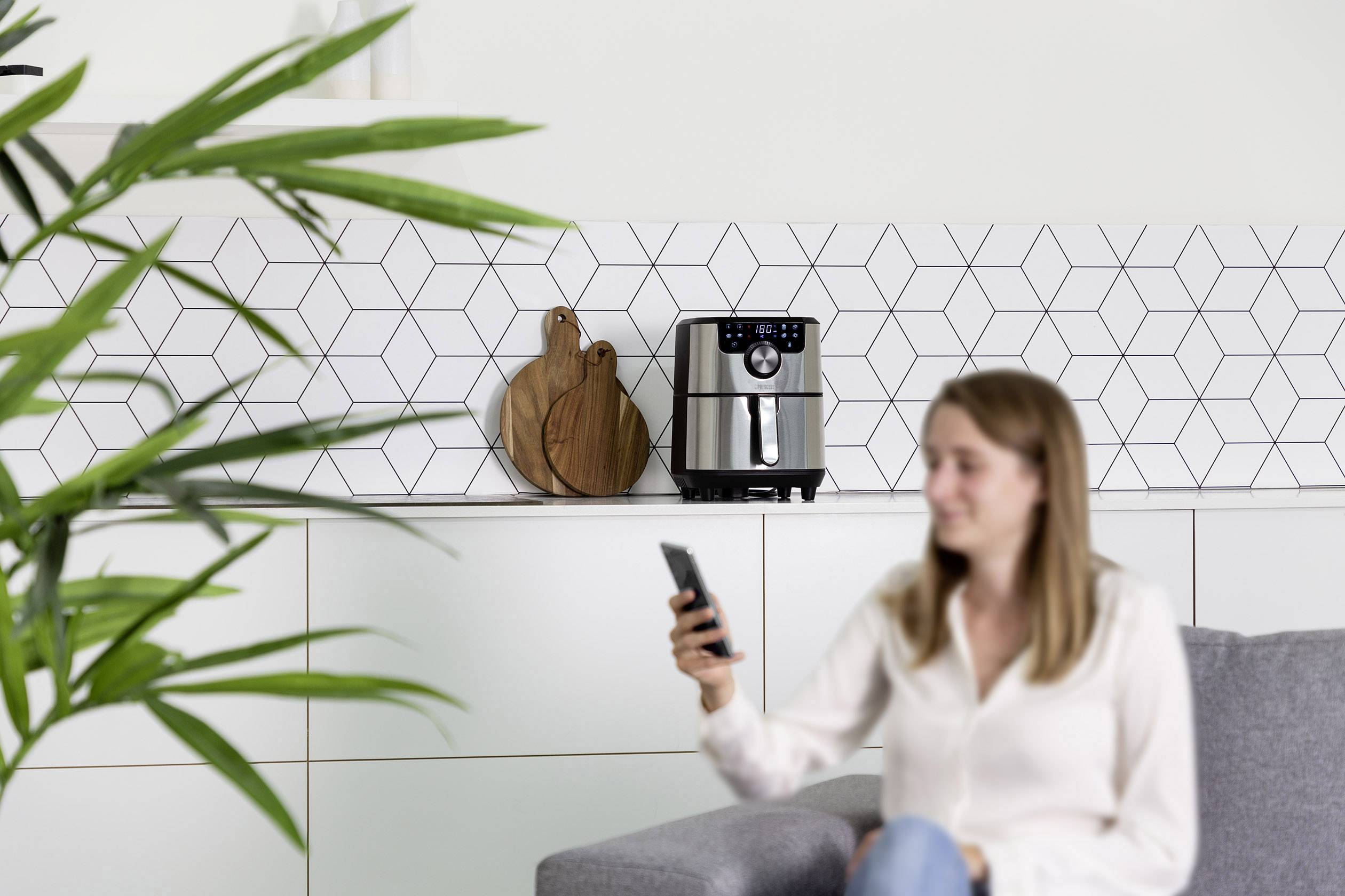 Eine Frau sitzt auf einem Sofa, hält ein Smartphone und schaut auf eine Heißluftfritteuse auf der Küchenarbeitsplatte im Hintergrund.