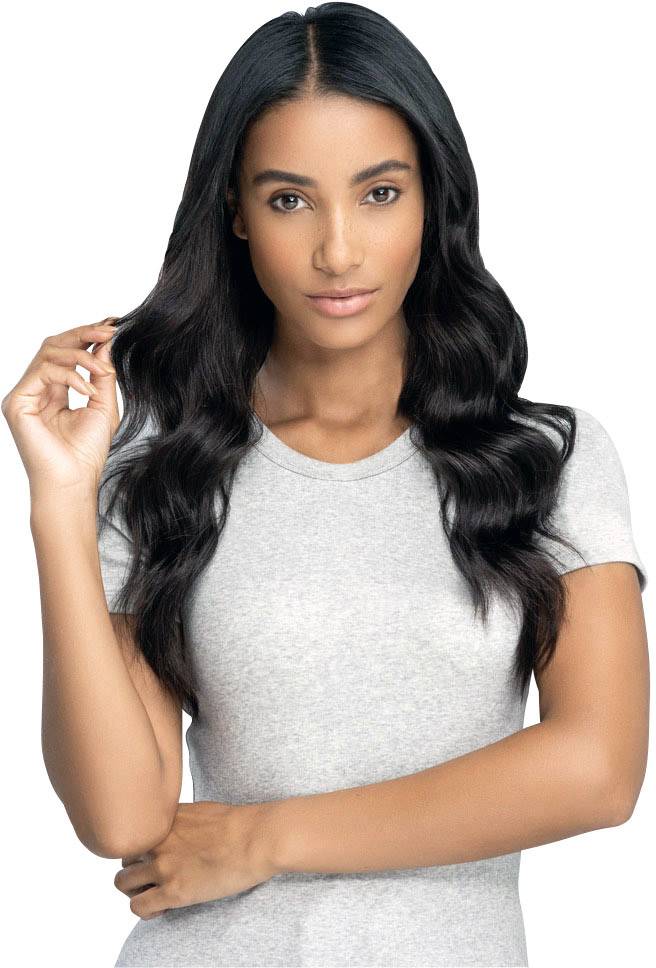 Eine Person mit langen, welligen Haaren und einem grauen T-Shirt schaut direkt in die Kamera, eine Hand im Haar.