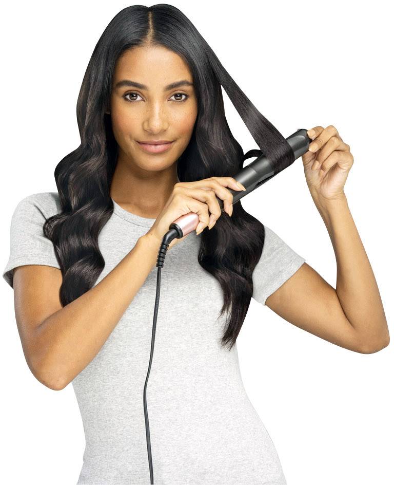 Eine Frau mit langen, gewellten Haaren benutzt ein Lockenstab, um ihre Haare zu stylen. Sie trägt ein graues T-Shirt.