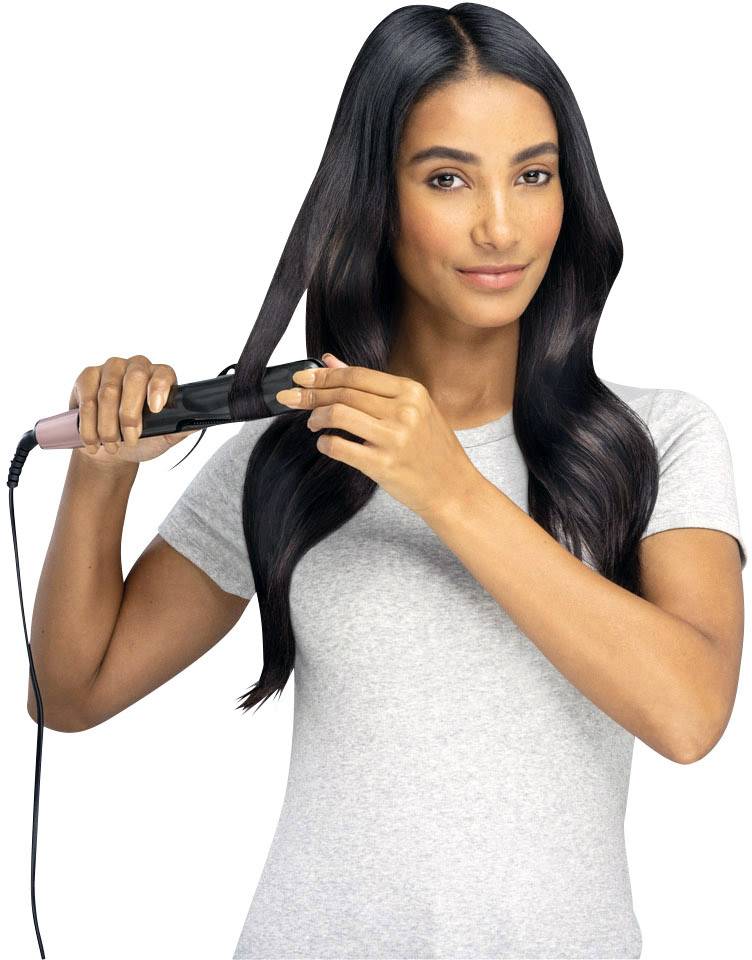 Frau mit langem, dunklem Haar glättet eine Haarsträhne mit einem Haarglätter, trägt ein graues T-Shirt.