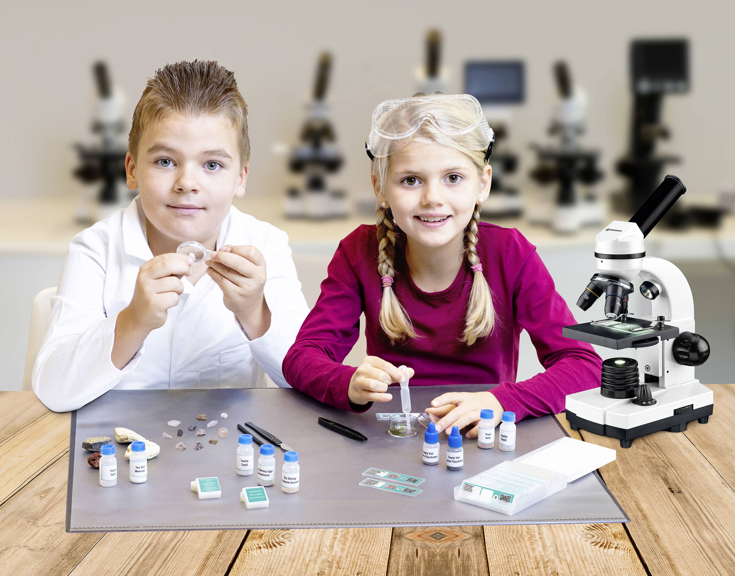 Kinder im Labor: Ein Junge und ein Mädchen untersuchen Proben mit einem Mikroskop. Sie tragen Schutzbrillen und teilen die Aufgabe.