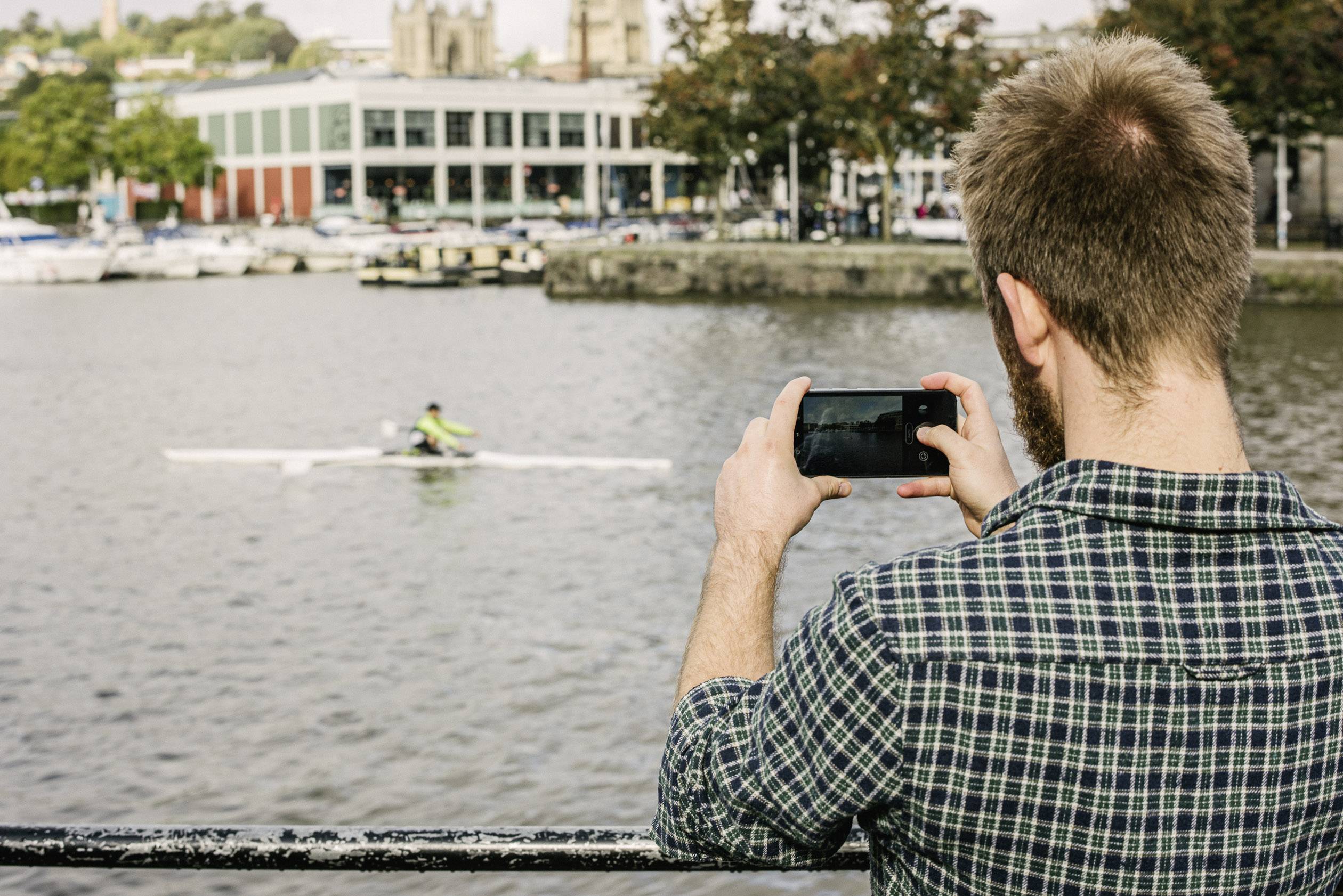 Ein Mann fotografiert mit seinem Smartphone einen Ruderer auf einem Fluss. Im Hintergrund ist ein Gebäude und Boote zu sehen.