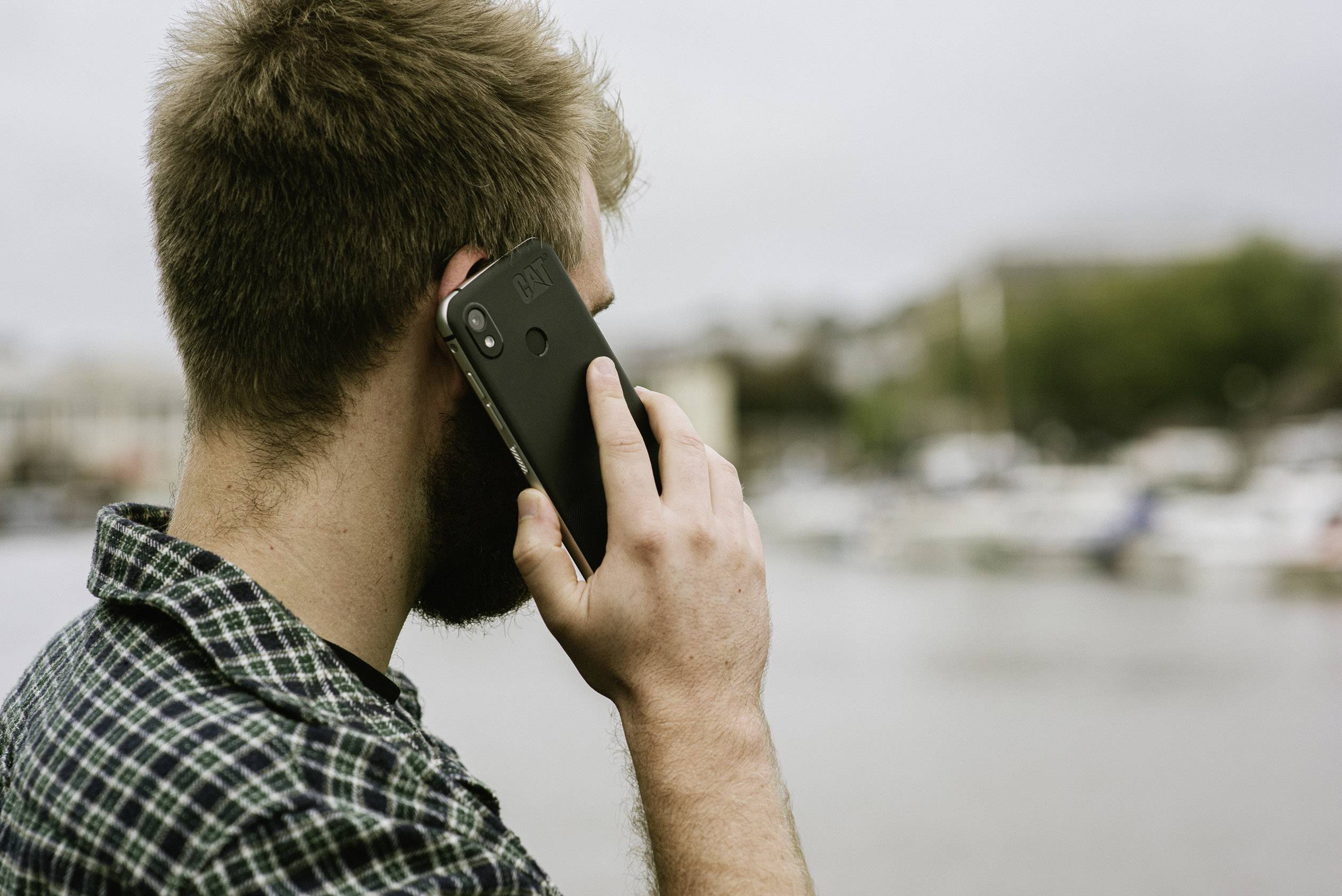 Eine Person telefoniert am Wasser mit einem Smartphone. Im Hintergrund sind unscharfe Boote und eine Stadt zu sehen.