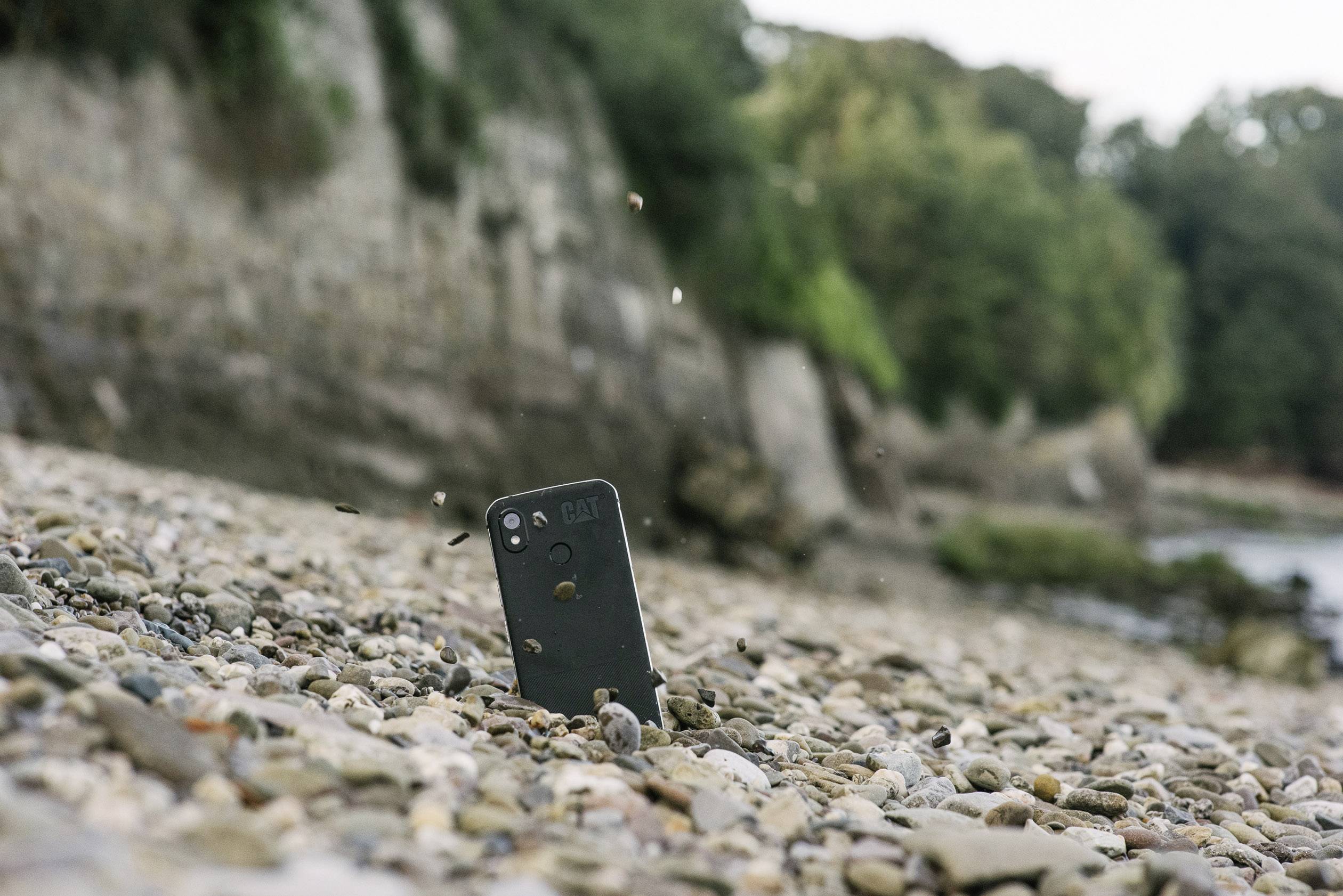 Ein Smartphone steht stabil in einem steinigen Uferbereich, umgeben von Kieselsteinen und bewaldeter Landschaft im Hintergrund.