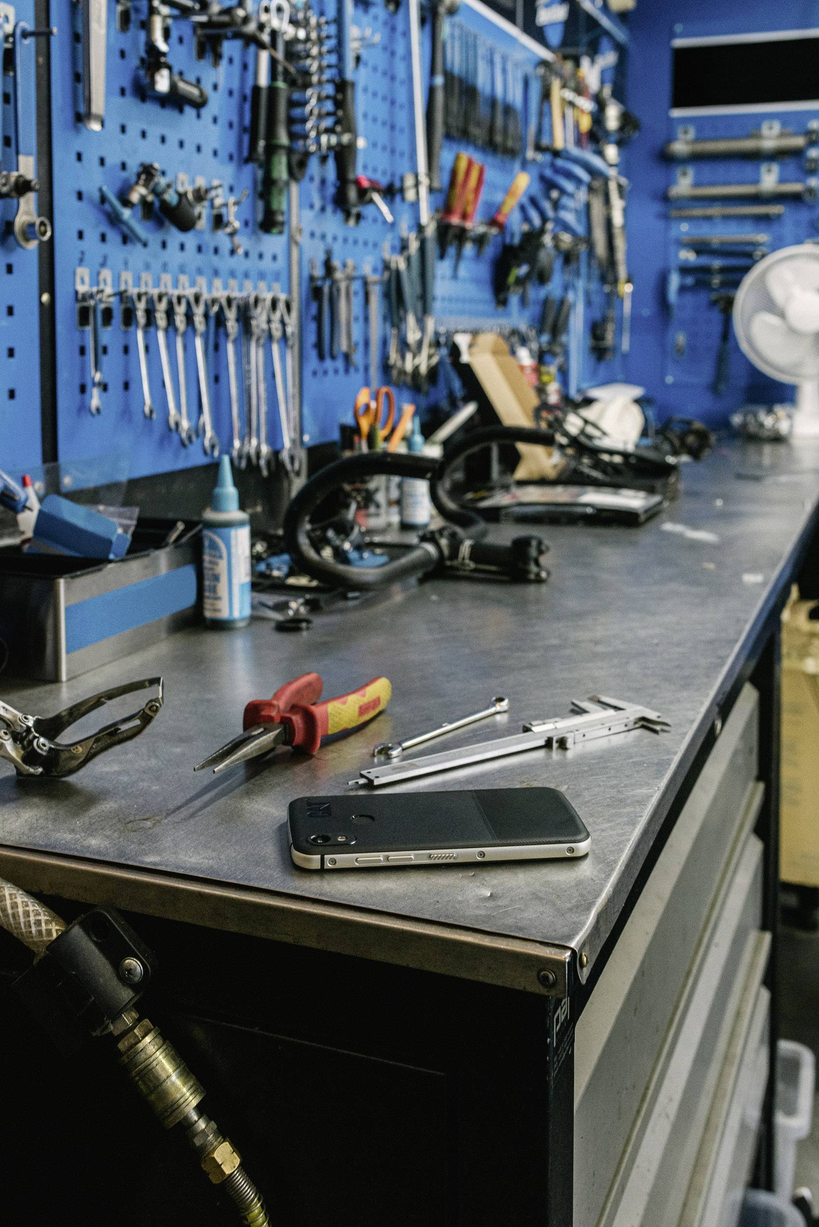 Eine Werkstatt mit einem Tisch voller Werkzeuge und Teilen. Ein Schraubenzieher, ein Lineal und ein Smartphone liegen auf dem Tisch. Im Hintergrund eine blaue Werkzeugwand.
