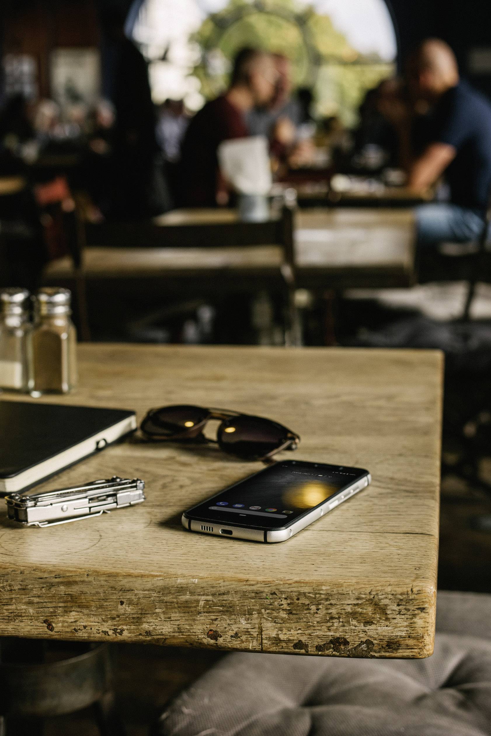 Ein Smartphone, Sonnenbrille und Notizbuch liegen auf einem Holztisch in einem Café. Im Hintergrund sind unscharfe Menschen erkennbar.