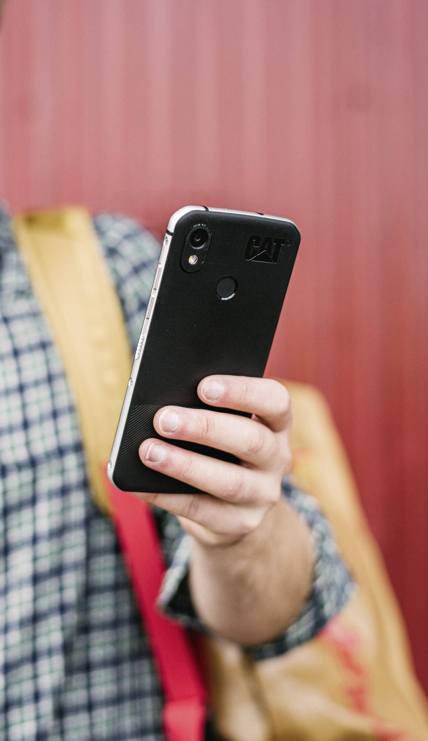 Eine Person hält ein Smartphone in der Hand, das sie mit dem Daumen bedient. Im Hintergrund trägt die Person einen Rucksack.