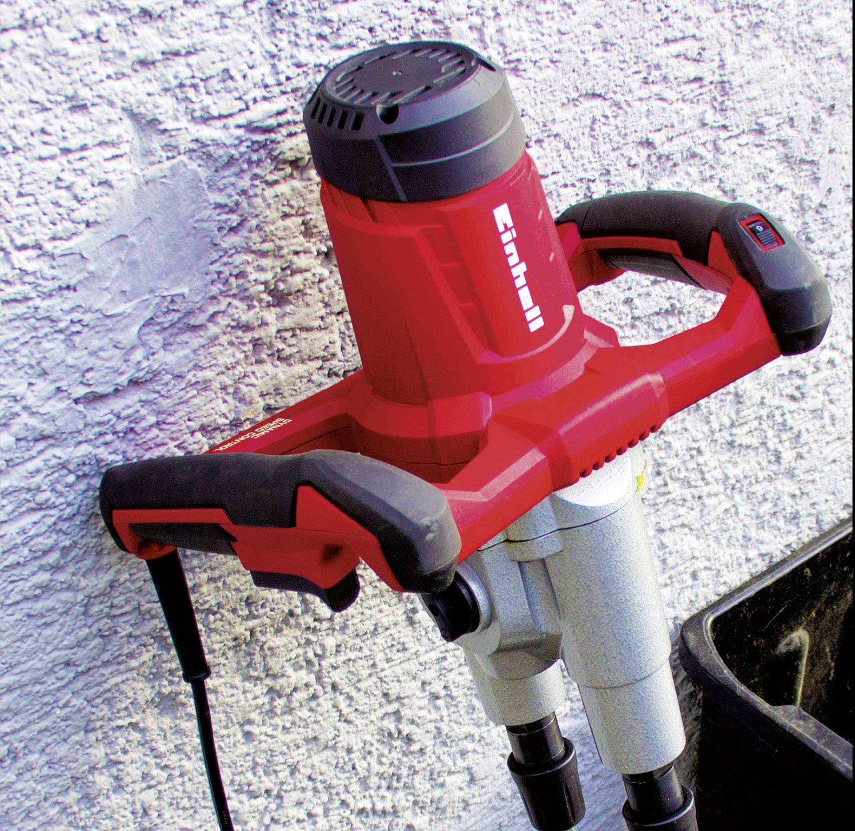 Ein roter Betonmischer ist an einer Wand befestigt. Das Gerät hat zwei Griffe und ein Stromkabel, das nach unten führt.