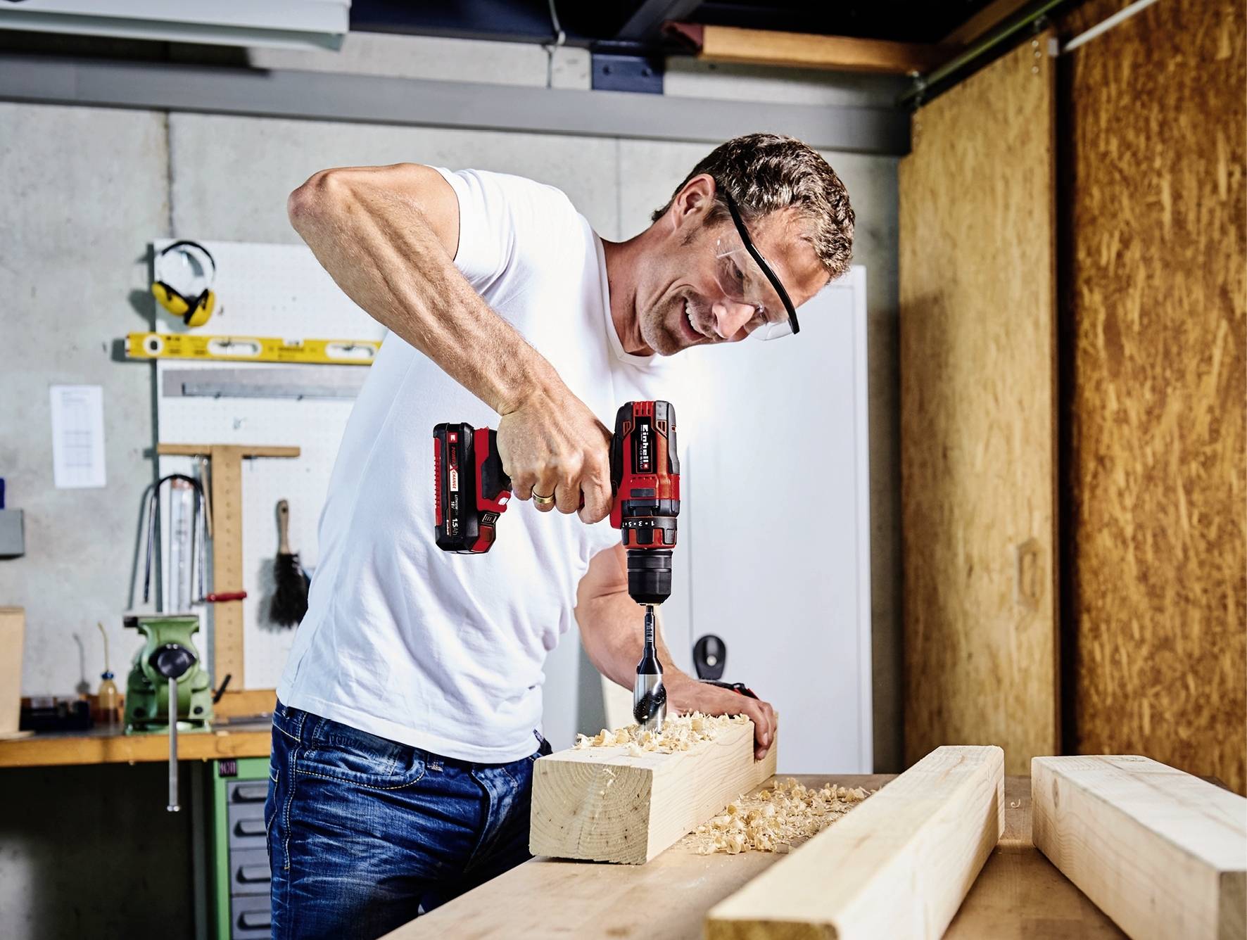 Ein Mann in einer Werkstatt bohrt mit einer roten Bohrmaschine ein Loch in einen Holzblock. Er trägt eine Schutzbrille und lächelt.