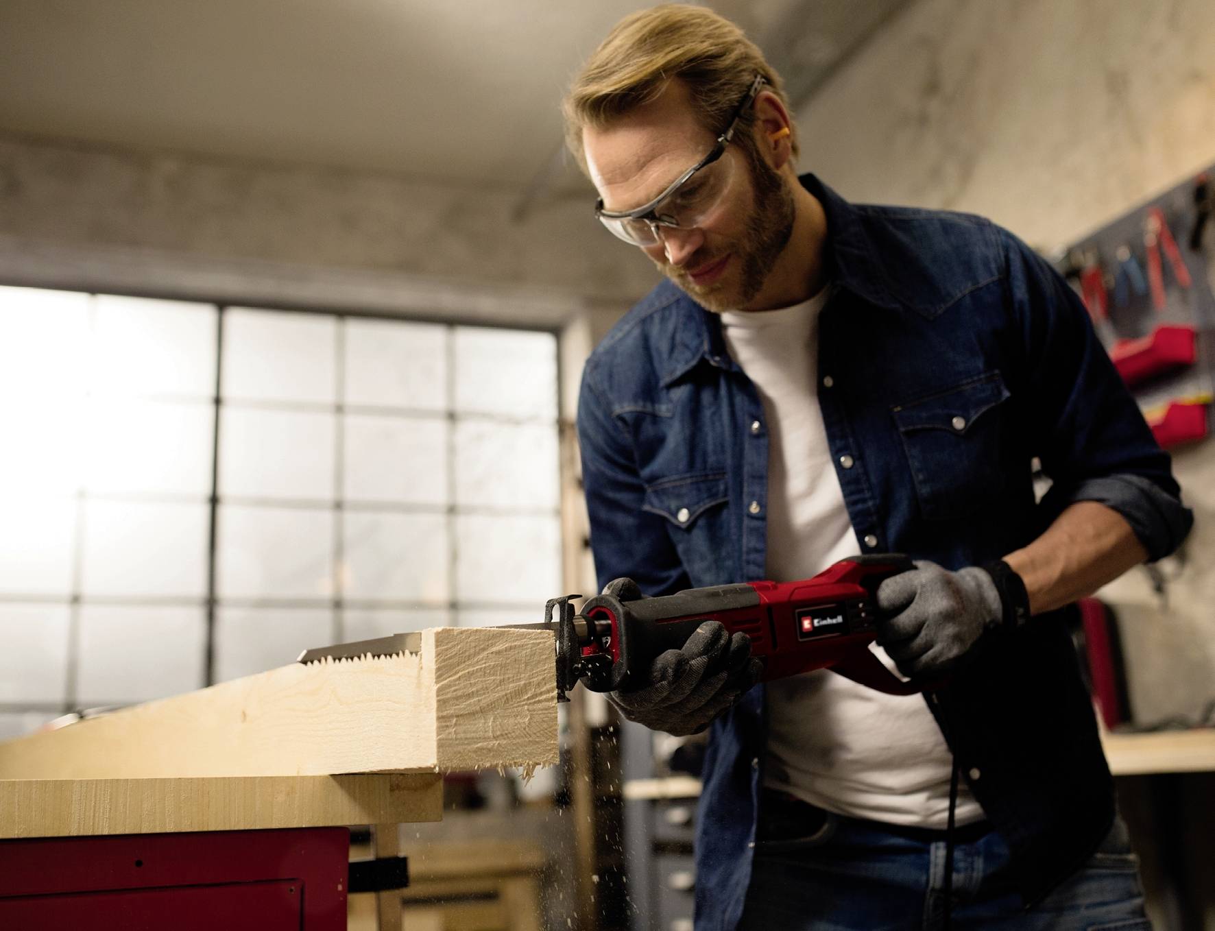Ein Mann in Schutzbrille und Arbeitshandschuhen sägt ein Holzbrett in einer Werkstatt mit einer elektrischen Säge.