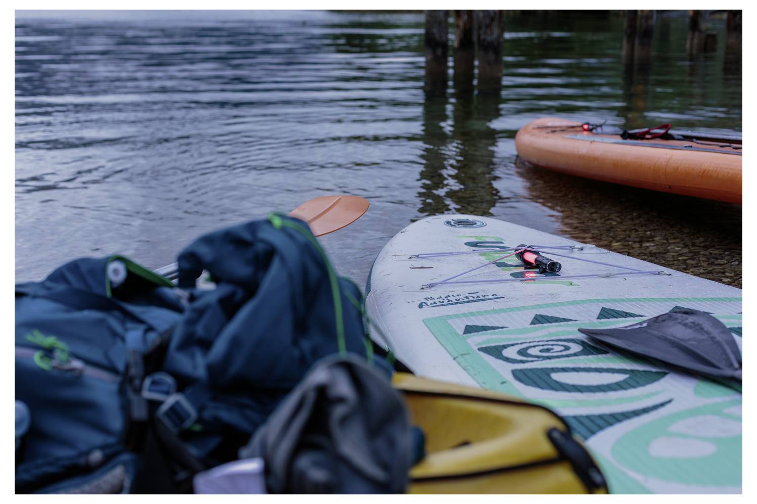 Stand-up-Paddleboards mit Taschen und Ausrüstung, die am Ufer eines ruhigen Sees oder Flusses ruhen und einsatzbereit sind.