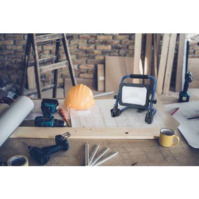 Arbeitsbereich in einer Werkstatt mit Skizzen, Helm, Werkzeugen, einer kleinen Lampe und einer Kaffeetasse auf einem Holztisch.