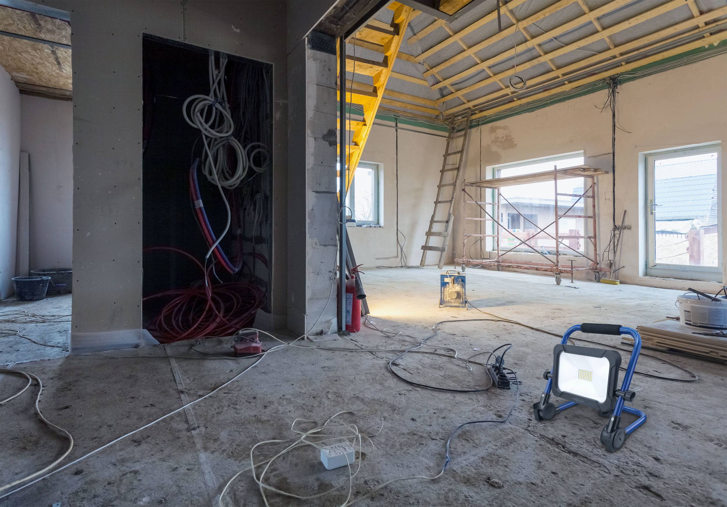 Eine Baustelle im Innenraum mit unfertigem Dach, Gerüst und barem Betonboden. Kabel hängen von der Decke, und eine Arbeitslampe steht am Boden.