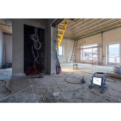 Eine Baustelle im Innenraum mit unfertigem Dach, Gerüst und barem Betonboden. Kabel hängen von der Decke, und eine Arbeitslampe steht am Boden.