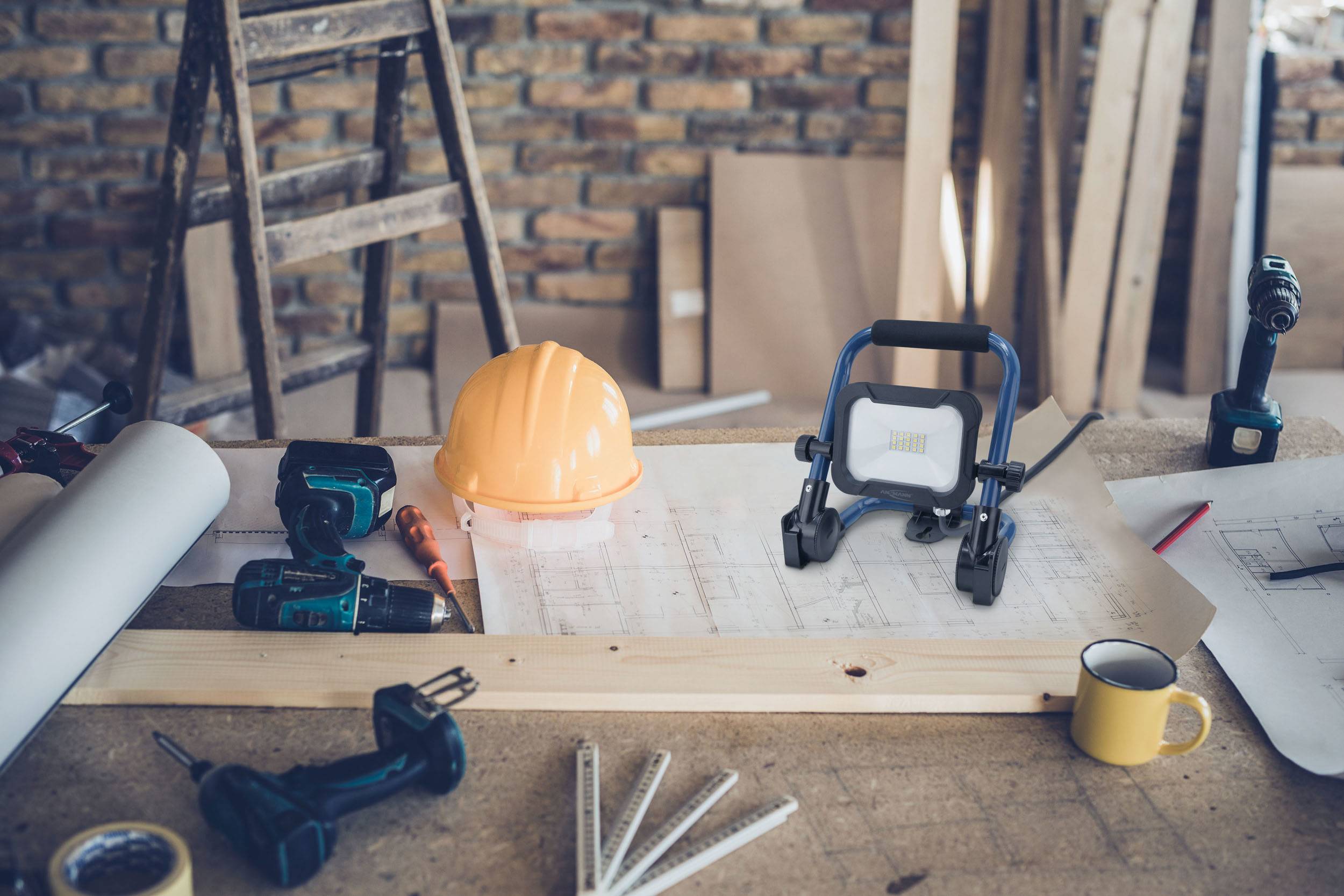 Arbeitsfläche in einer Werkstatt mit Bauplänen, Helm, Werkzeugen und einem Baustrahler, die eine aktive Bauumgebung darstellen.