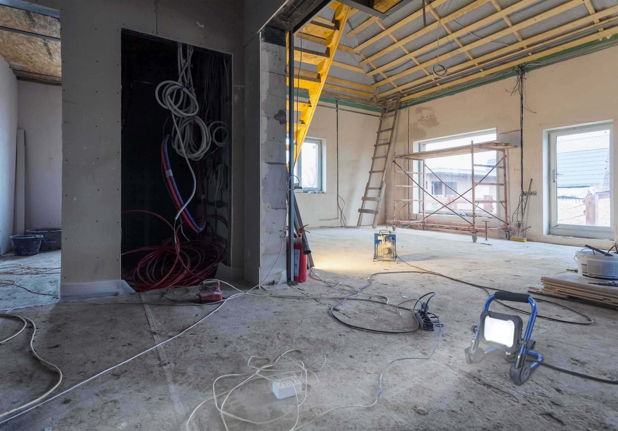 Innenansicht einer Baustelle mit unfertigem Dach, freiliegenden Kabeln und Baugerüsten. Ein Baugerüst steht neben den großen Fenstern.