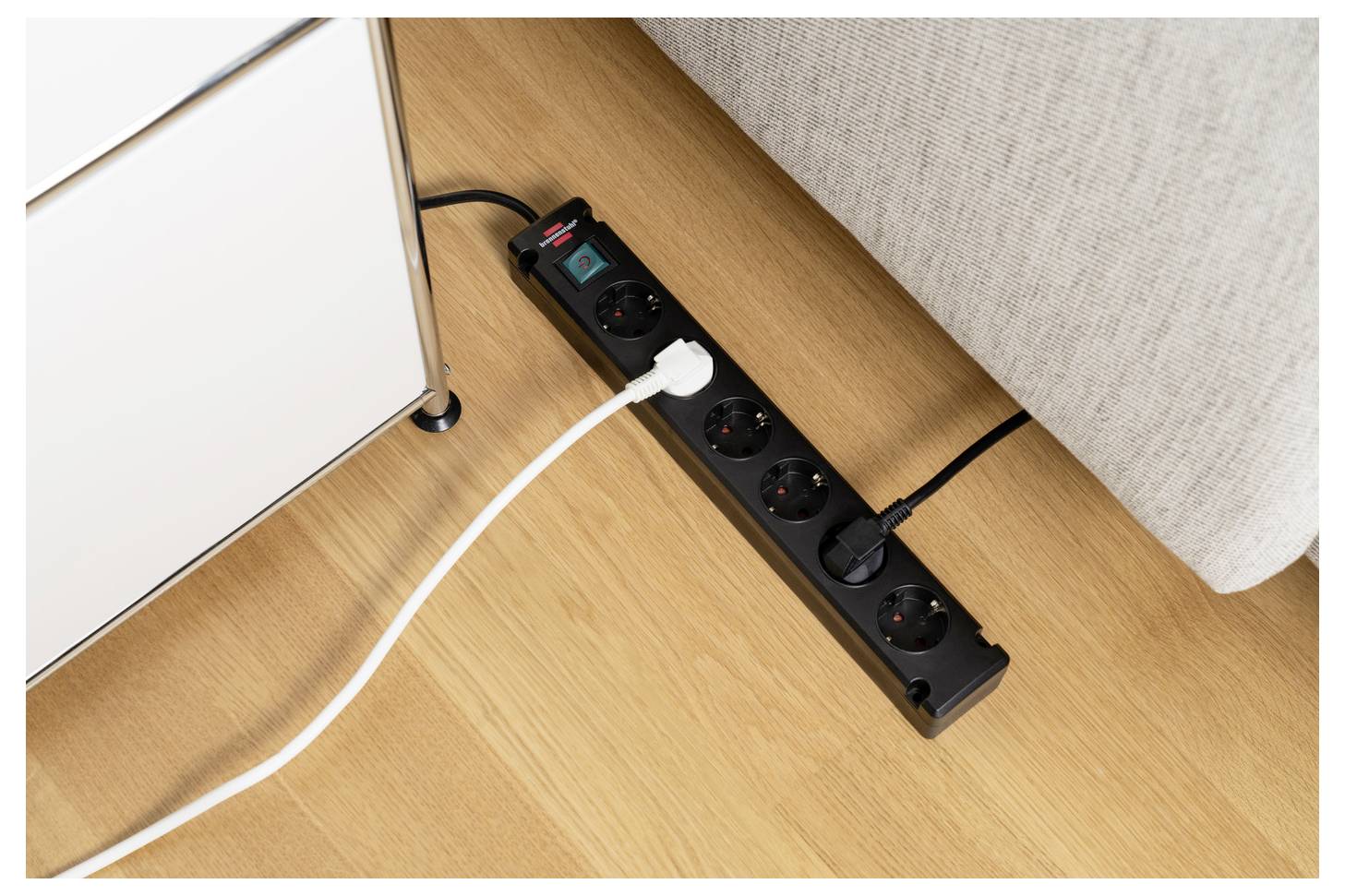 Schwarze Steckerleiste auf einem Holzboden neben Möbeln, mit mehreren Steckern und Kabeln verbunden, die eine elektrische Einrichtung auf Bodenniveau zeigen.