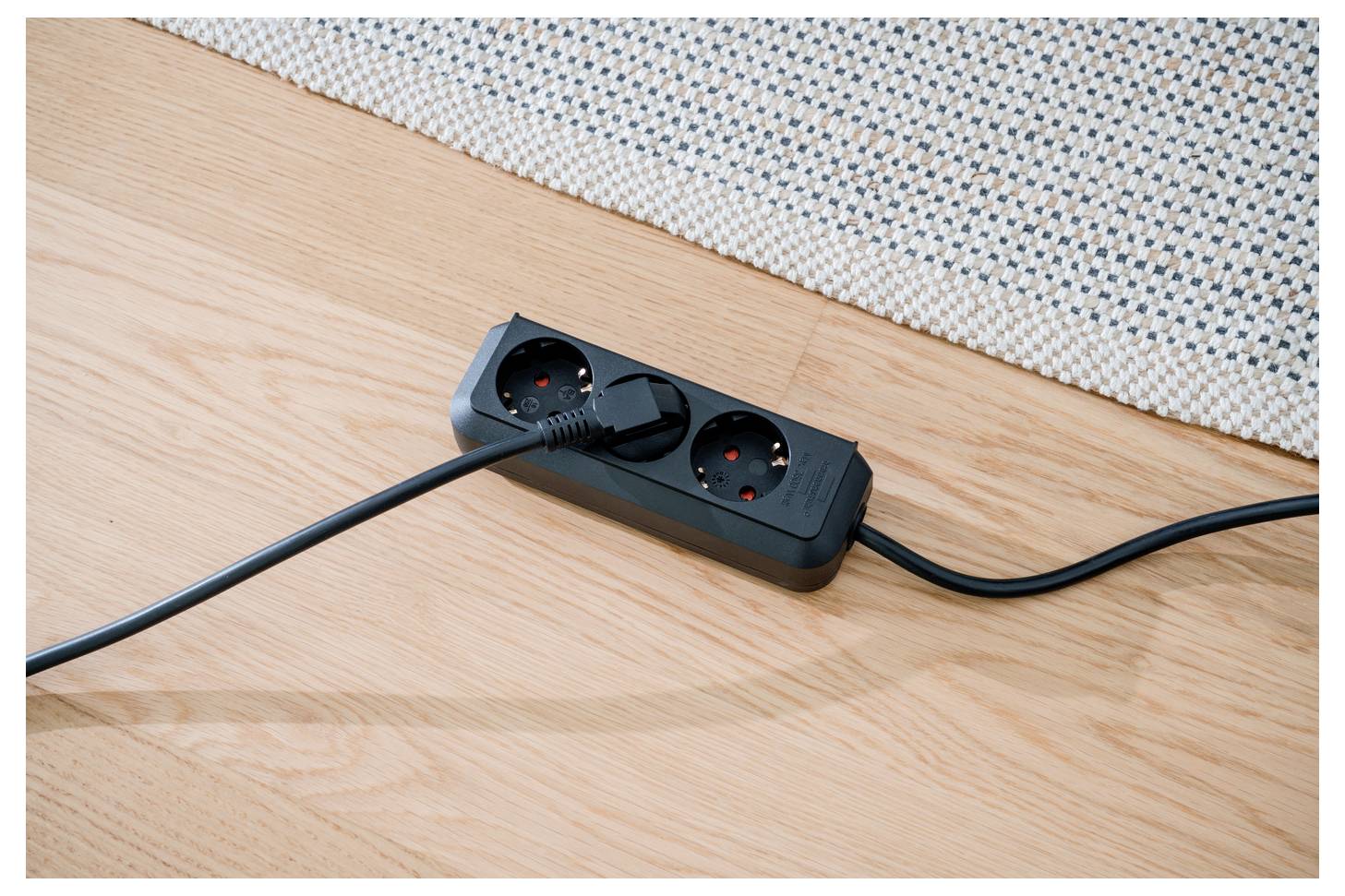 Schwarze Steckerleiste mit zwei eingesteckten Kabeln auf einem Holzboden neben einem Teppich.