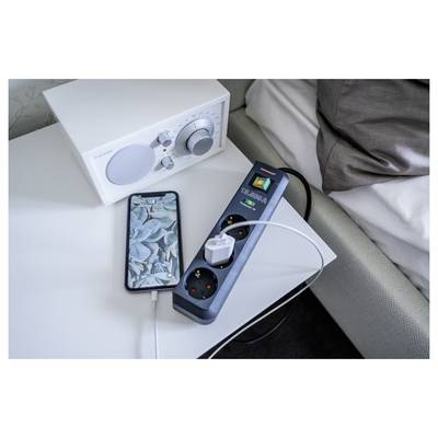 Smartphone-Ladegerät neben einem weißen Lautsprecher und einer Steckerleiste mit mehreren angeschlossenen Geräten auf einem Nachttisch.