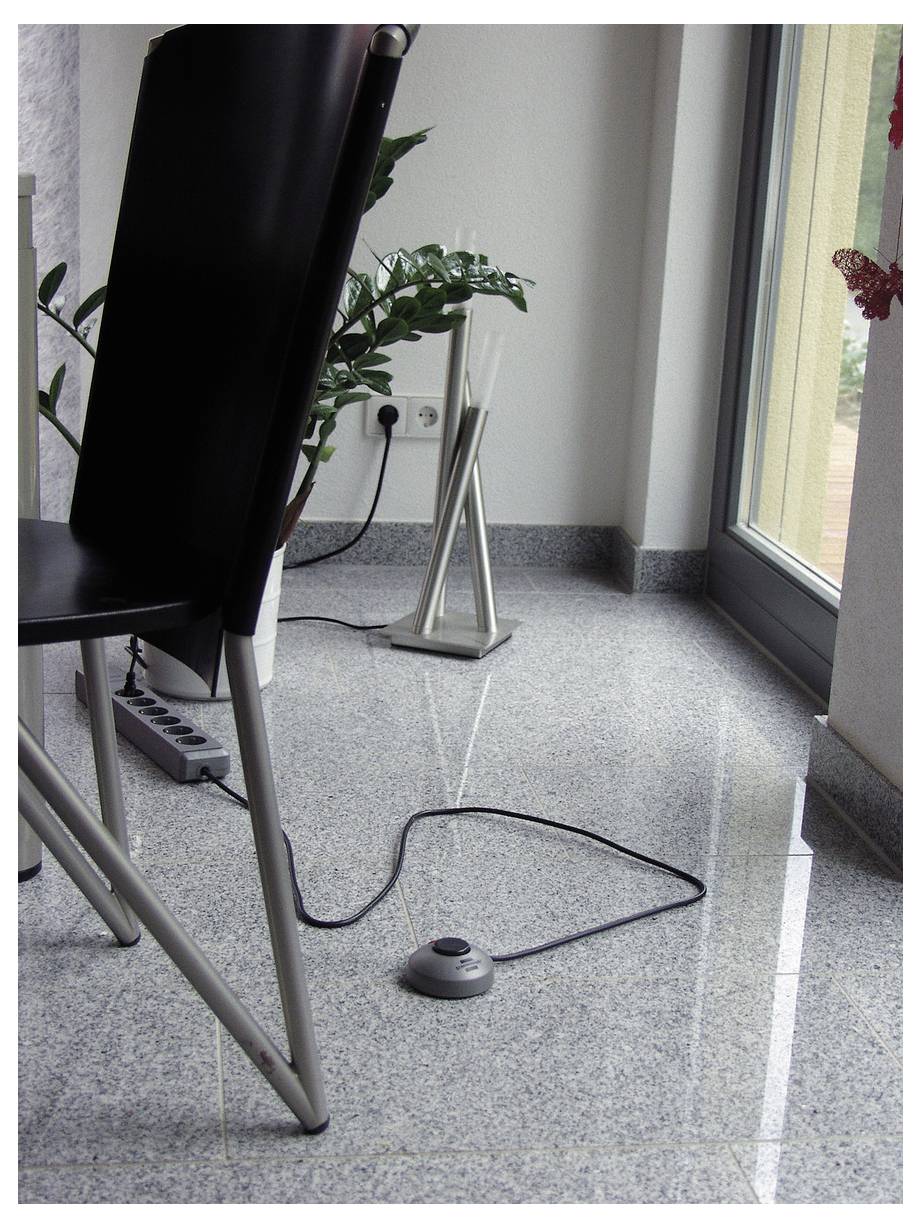 Ein verkabeltes Fußpedal auf einem gefliesten Boden neben einem Stuhl, in der Nähe einer Wandsteckdose und eines Fensters, was auf ein fußbetriebenes Steuergerät hindeutet.