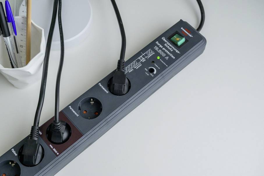 Eine Steckdosenleiste mit mehreren angeschlossenen Kabeln auf einem weißen Tisch. Ein Bein eines Lampenfußes und ein Behälter mit Stiften sind im Hintergrund.