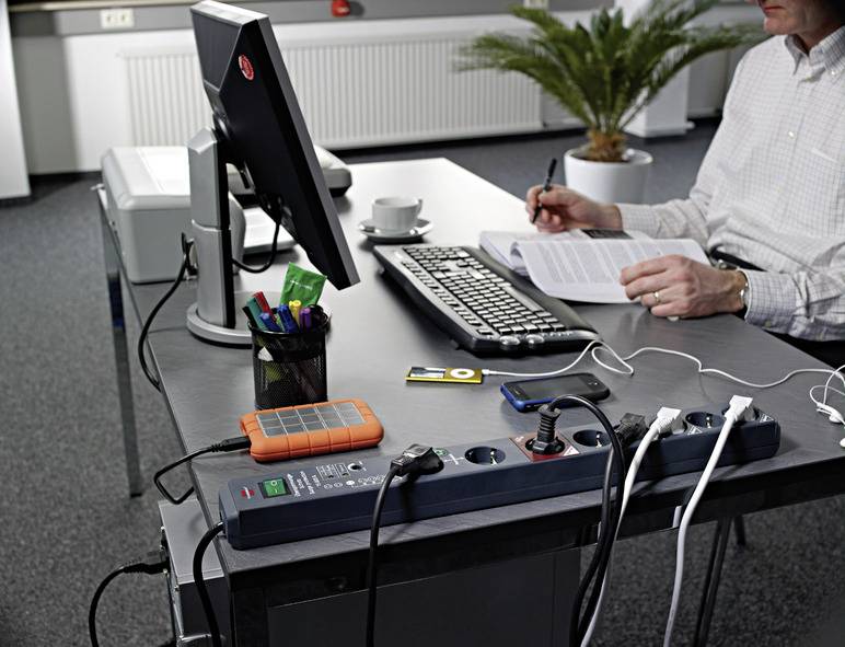 Eine Person sitzt an einem Schreibtisch mit Computer, Tastatur und Notizen. Mehrere Geräte sind an eine Steckdosenleiste angeschlossen.