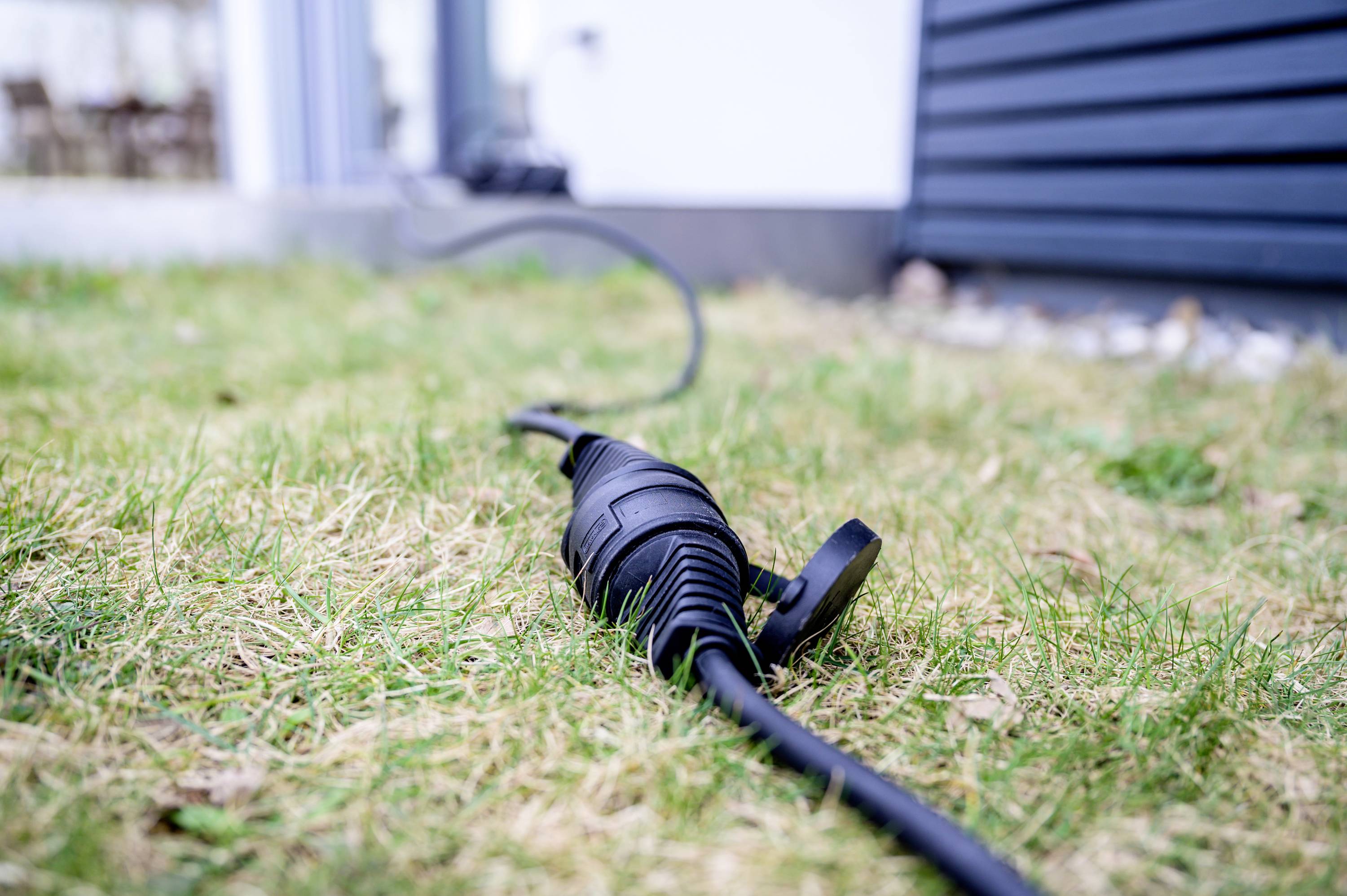 Nahaufnahme eines schwarzen Stromkabels auf grün-braunem Rasen, das zu einem Haus führt. Im Hintergrund ist ein Gebäude erkennbar.